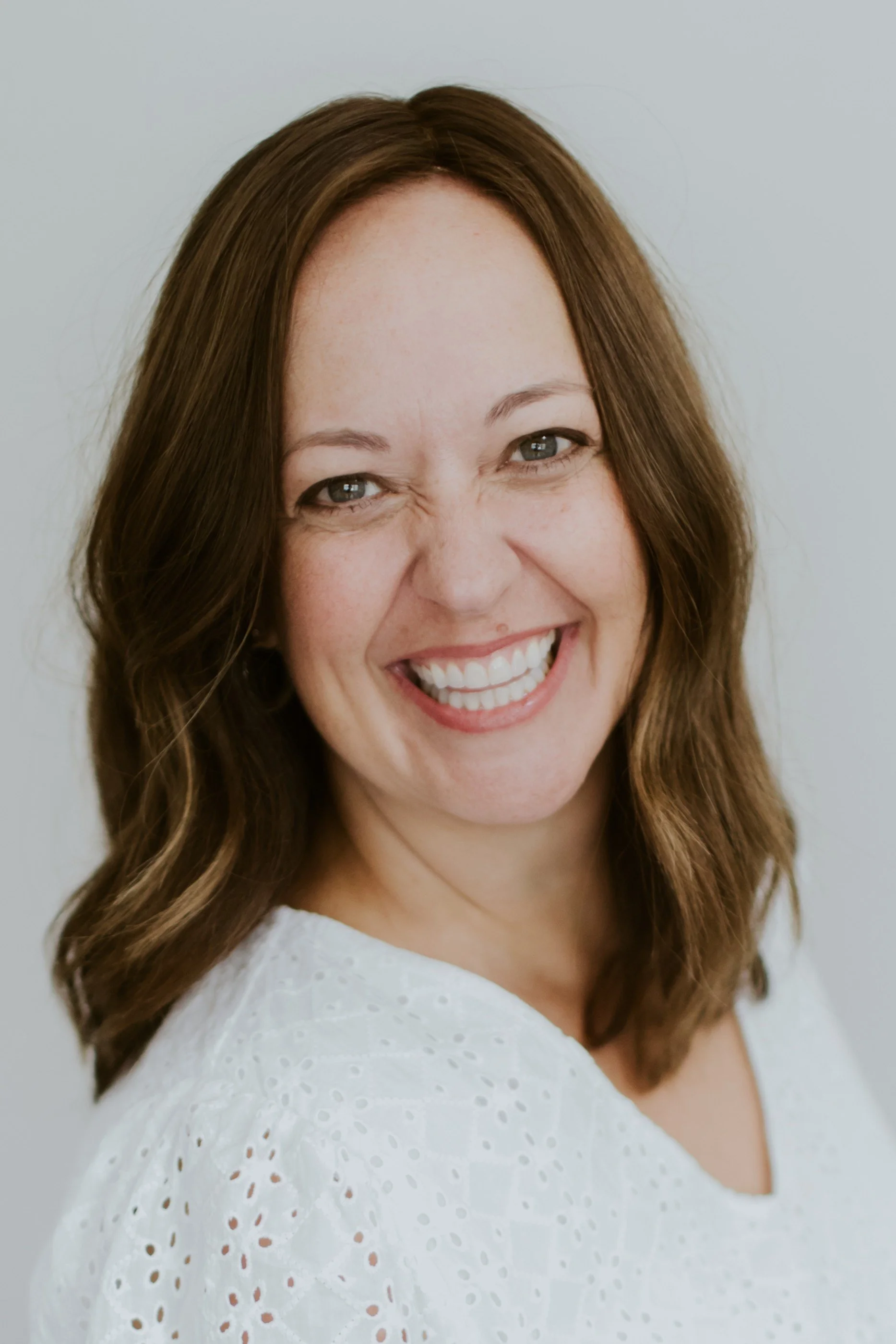 A woman with long, wavy brown hair and blue eyes, smiling joyfully, wearing a white eyelet top, standing against a plain light background.
