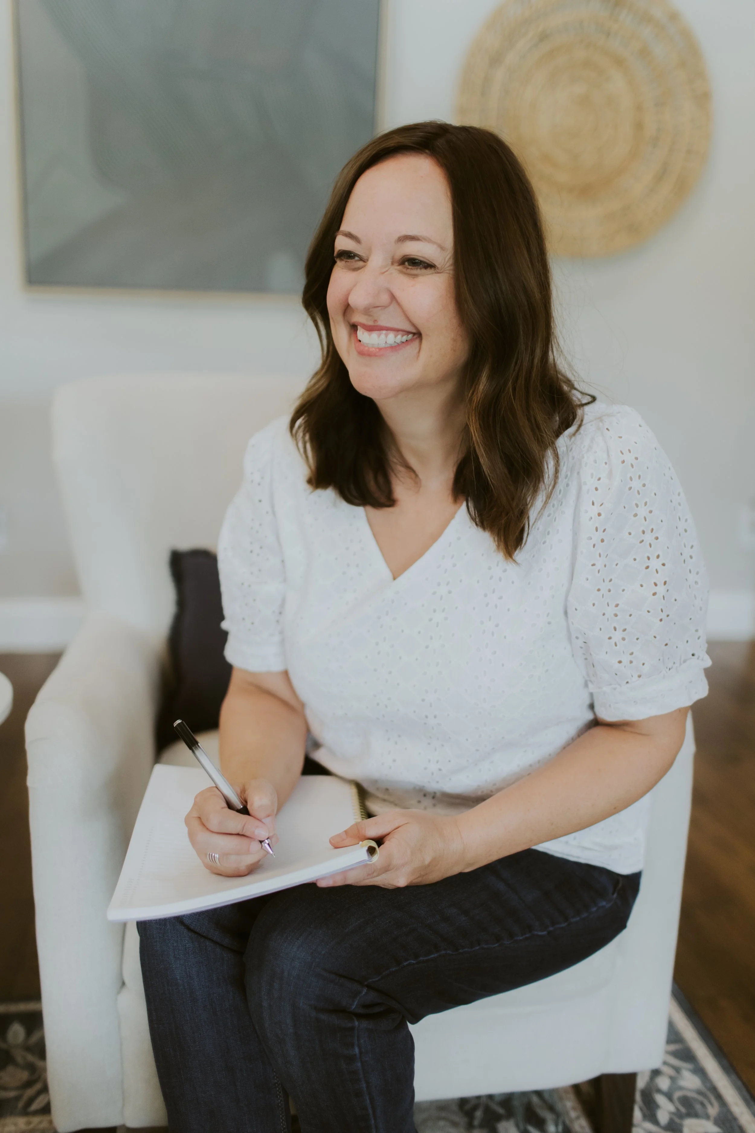 A woman with shoulder-length brown hair, smiling, sitting on a white armchair, holding a pen, and writing in a spiral notebook, in a room with white walls, a framed abstract artwork, and a woven wall decor.