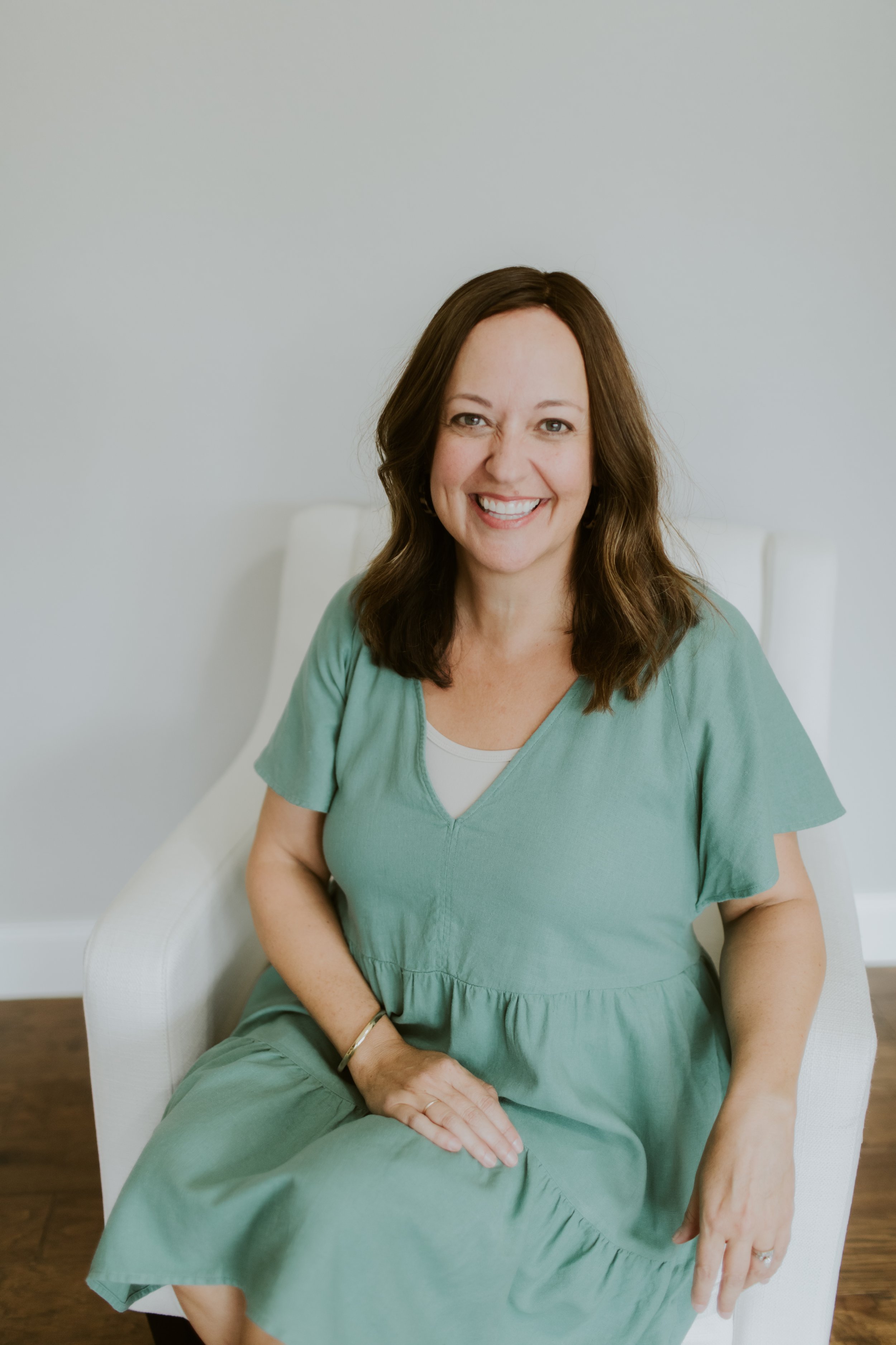 A smiling woman with long brown hair sitting in a white chair against a light gray wall, wearing a green dress.