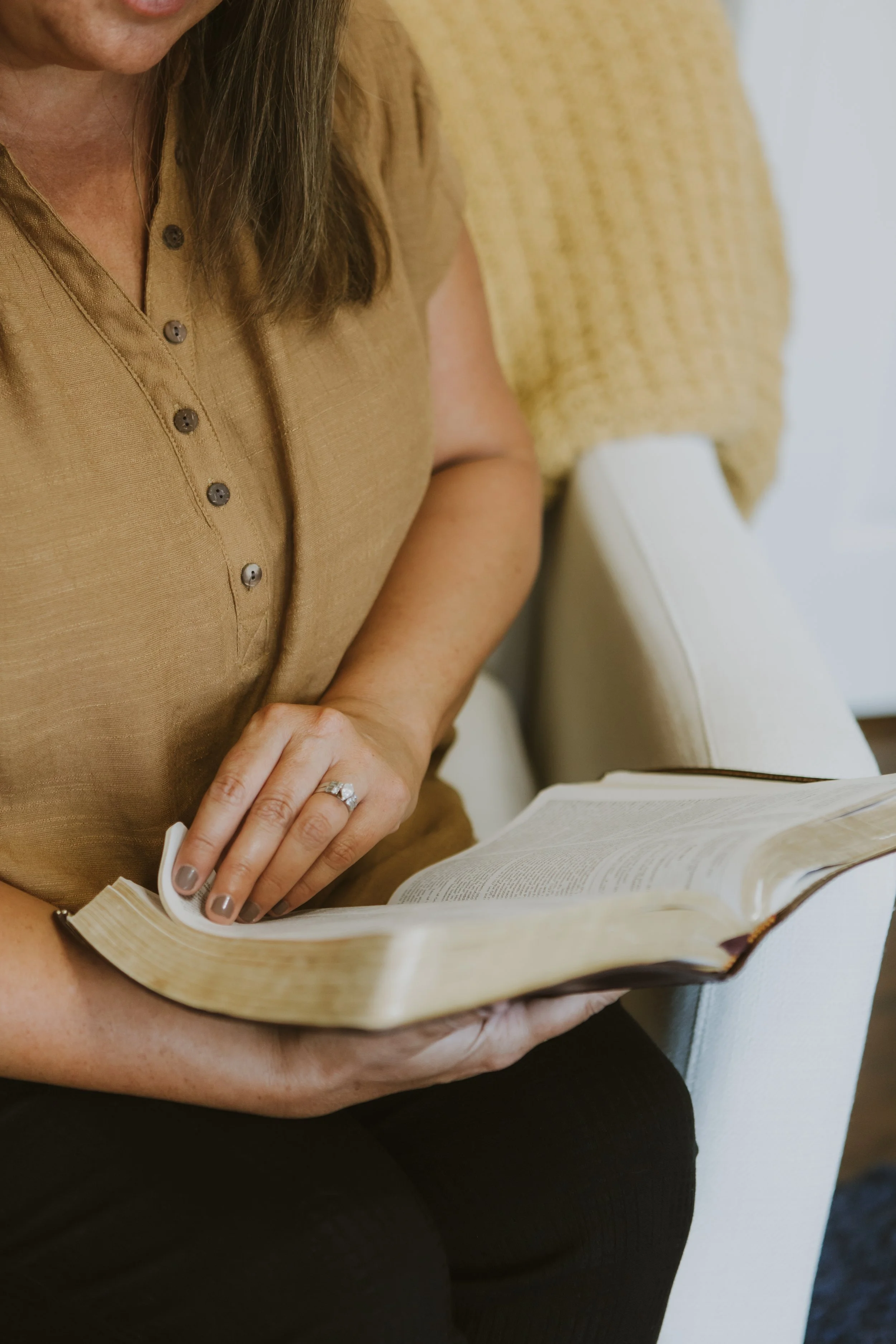 Close-up of a woman reading a book, wearing a tan short-sleeved shirt, sitting on a white chair.