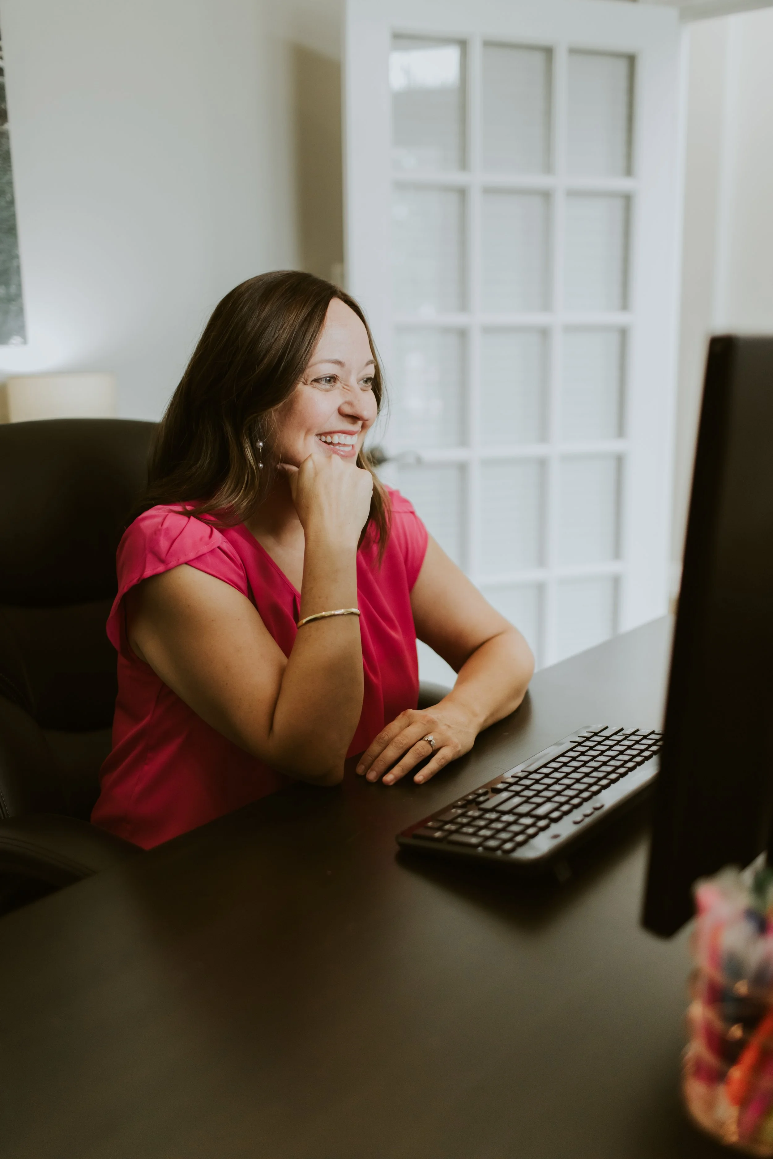 A woman with brown hair wearing a pink blouse is smiling and looking at a computer screen. She is sitting at a desk with a keyboard in front of her in a bright room with a large window in the background.
