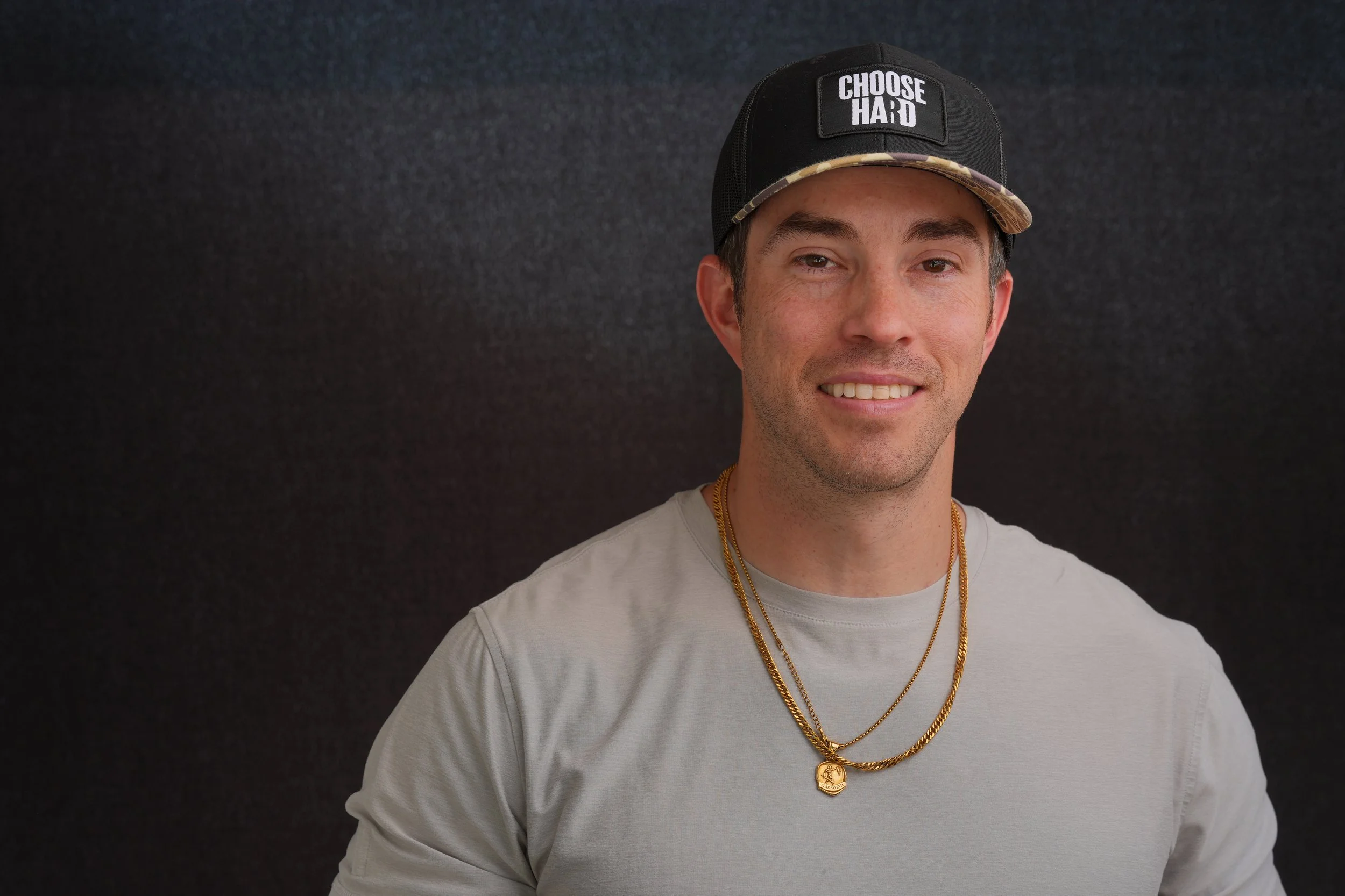 A young man smiling, wearing a black cap with the phrase 'CHOOSE HARD' on the front, layered gold necklaces, and a light-colored sweatshirt against a dark background.