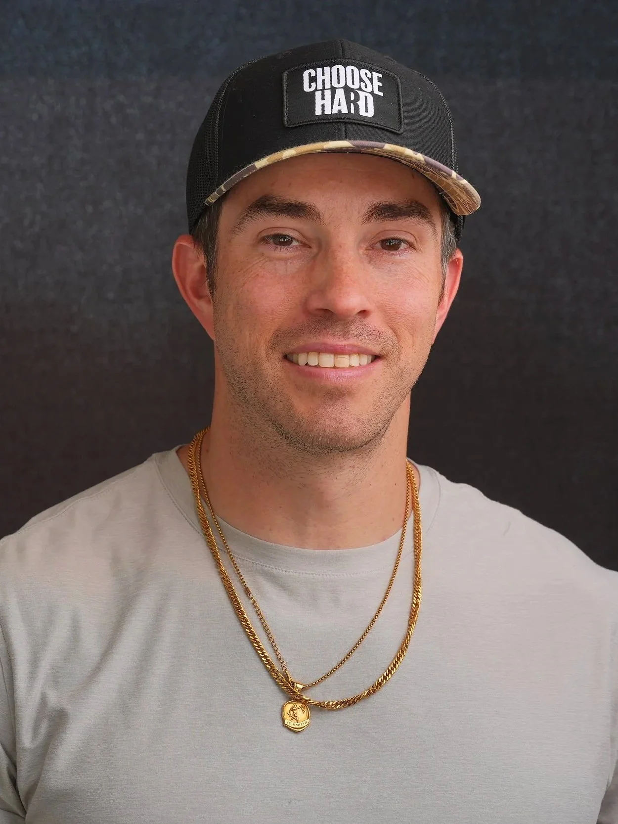 A man wearing a black baseball cap with the words 'CHOOSE HARD' on the front, a light gray t-shirt, and gold chains with a pendant, standing against a dark background.