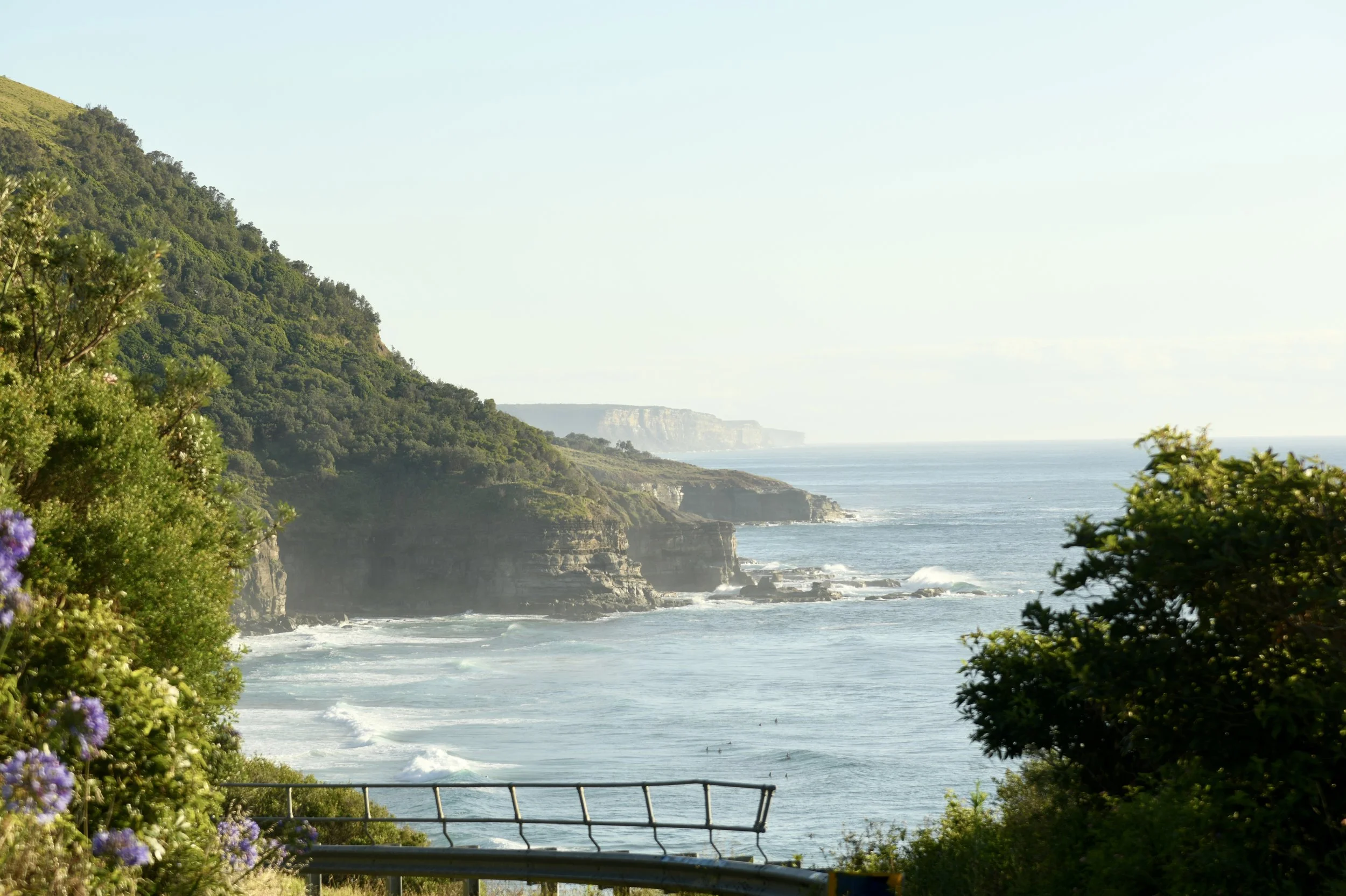 Coastal landscape with cliffs, ocean waves, and greenery in the foreground, under a clear sky.