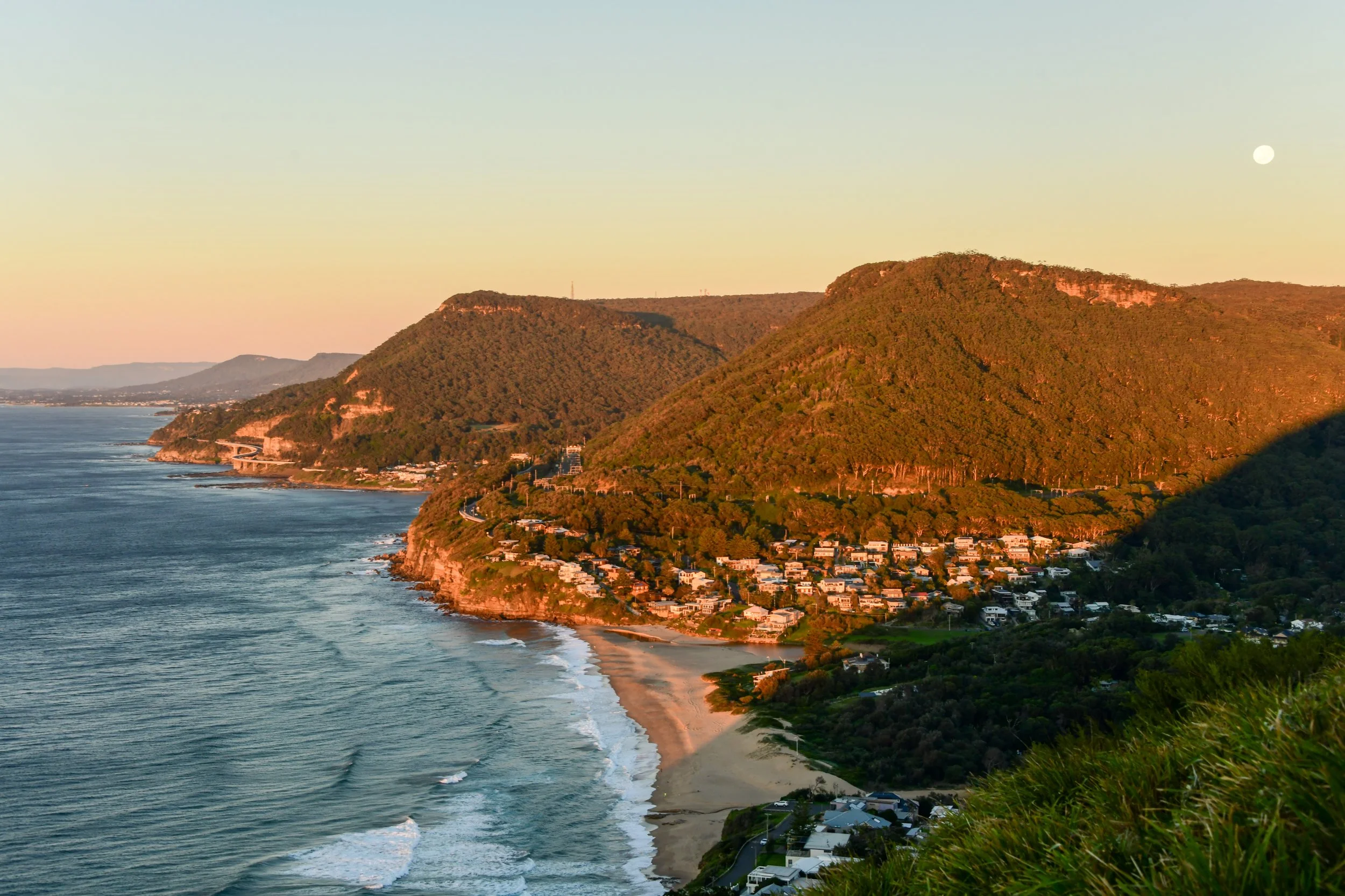 Sunset view of a coastal hillside with houses, a beach, and the ocean, with a full moon in the sky.
