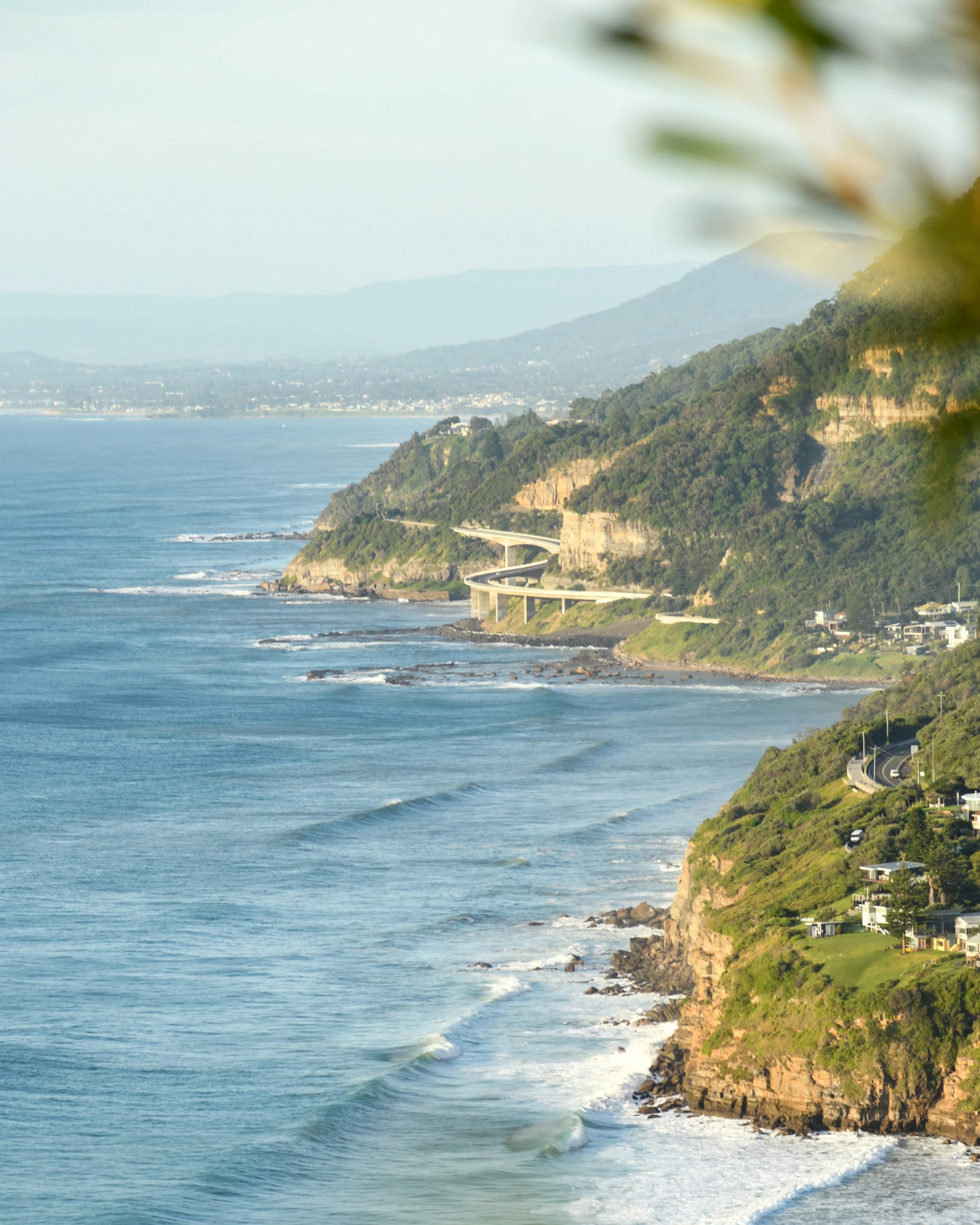 Coastal view of cliffs with scenic winding road, houses, green vegetation, and ocean waves, with distant mountains in the background.