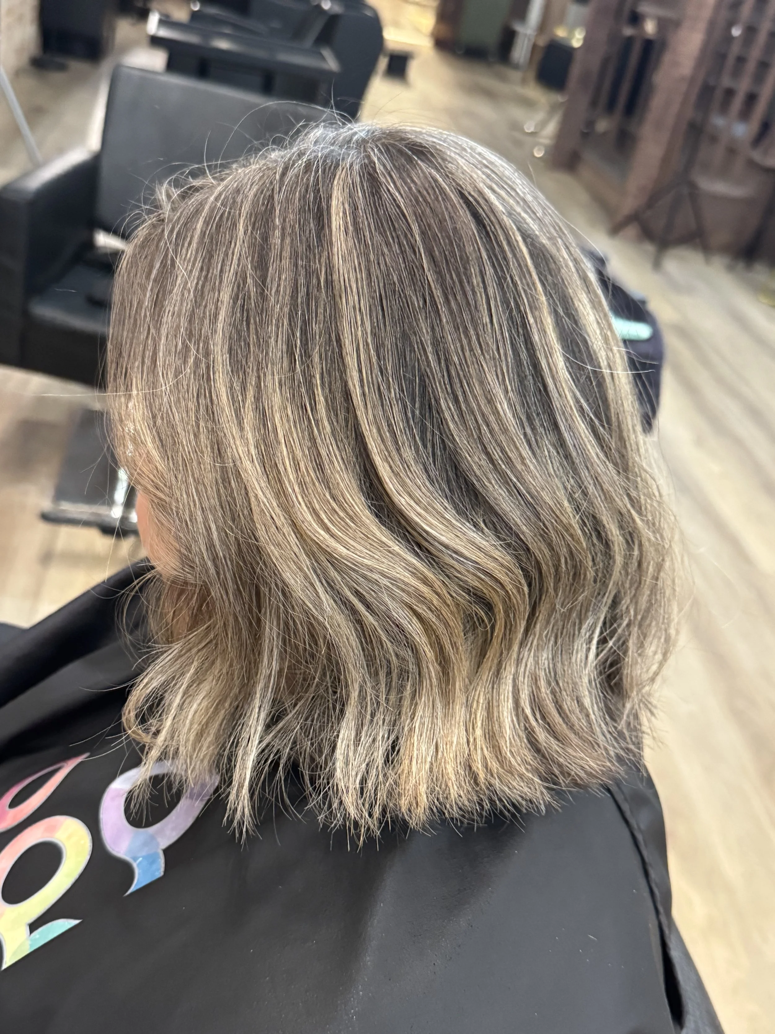 Back view of a woman's head with shoulder-length, blonde hair featuring highlights, sitting on a black chair in a salon with salon chairs and hair styling stations in the background.