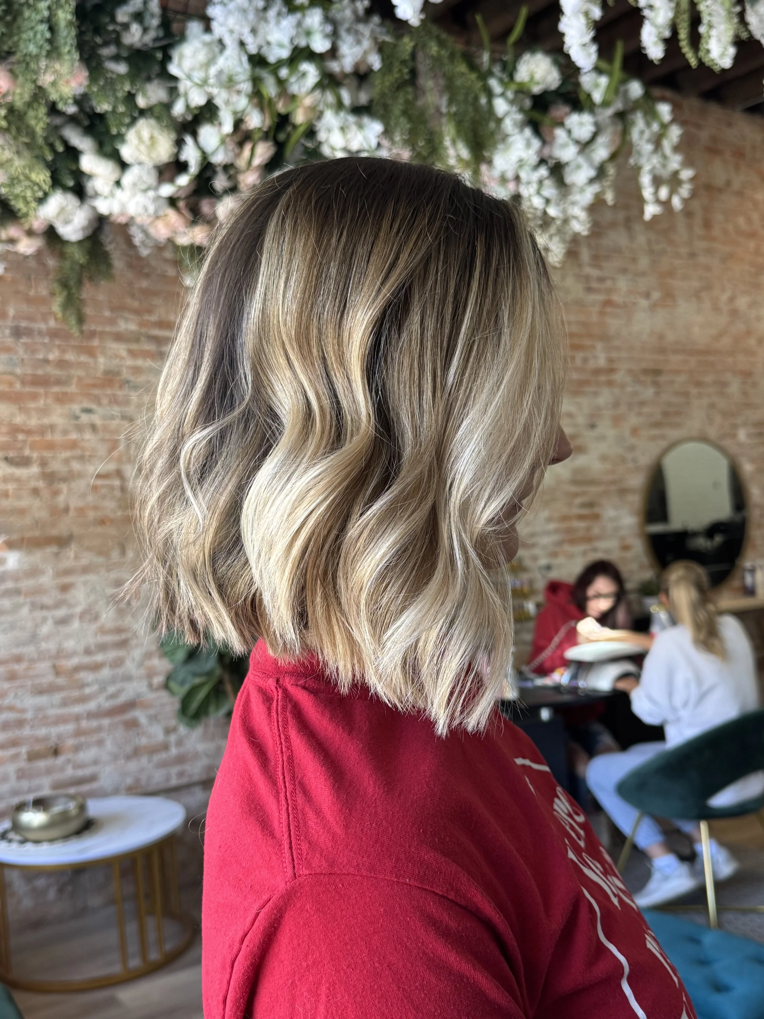 Side view of a woman with long, wavy blonde hair wearing a red sweatshirt in a cafe setting with a brick wall and people sitting at tables in the background.