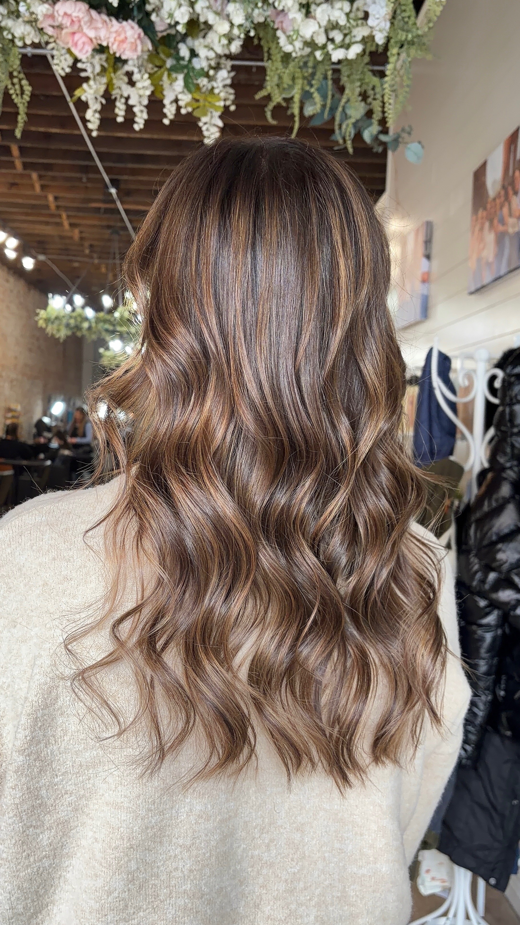 Back view of a woman with long, wavy, light brown hair with subtle highlights, indoors with floral decor hanging from the ceiling and people in the background.