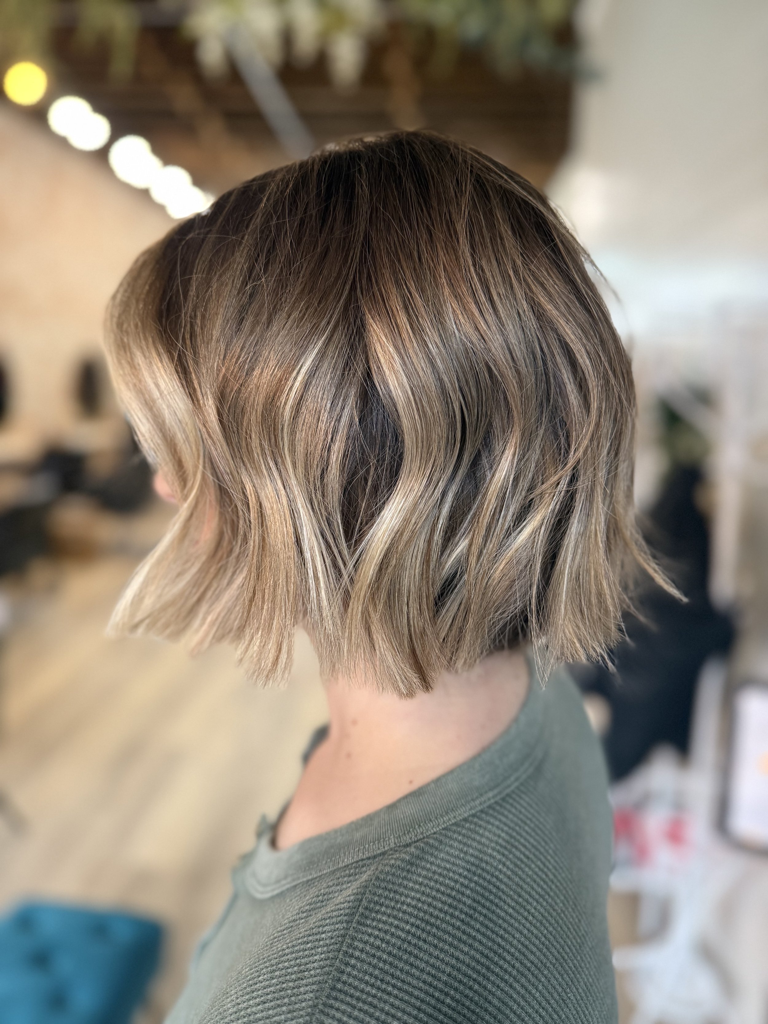 Back view of a person with short, styled, blonde hair with darker roots, wearing a green top.