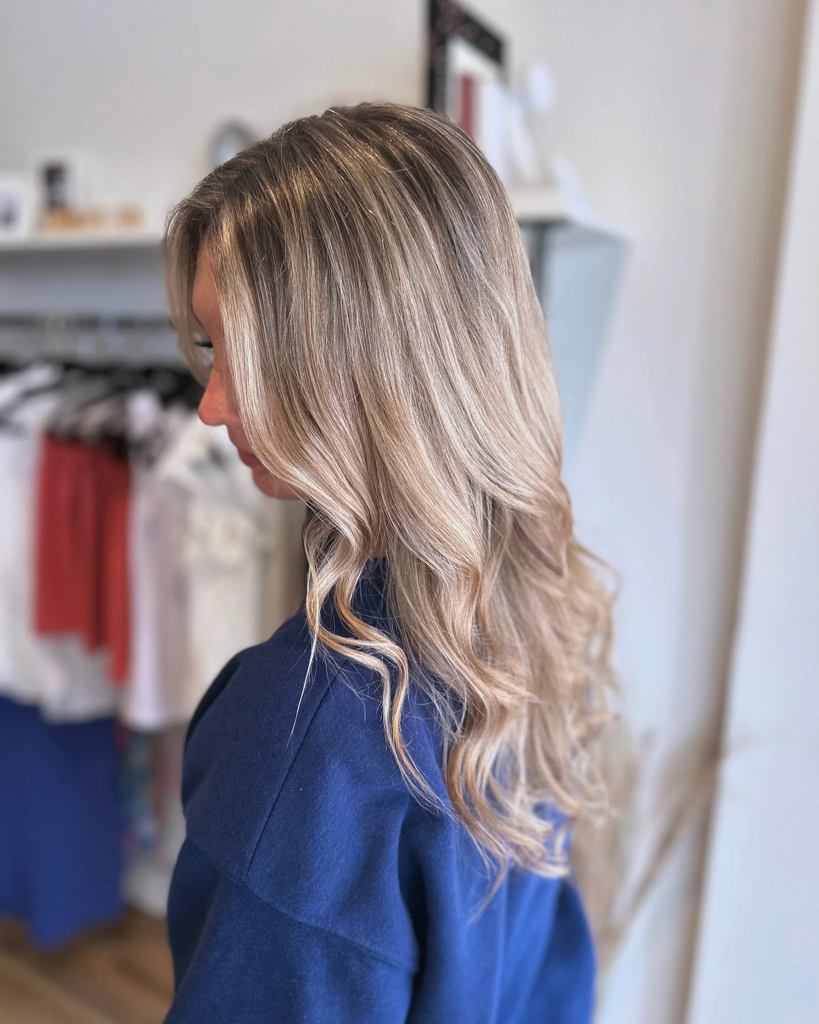 A woman with wavy, blonde hair styled in loose curls, wearing a navy blue top, indoors with blurred background.