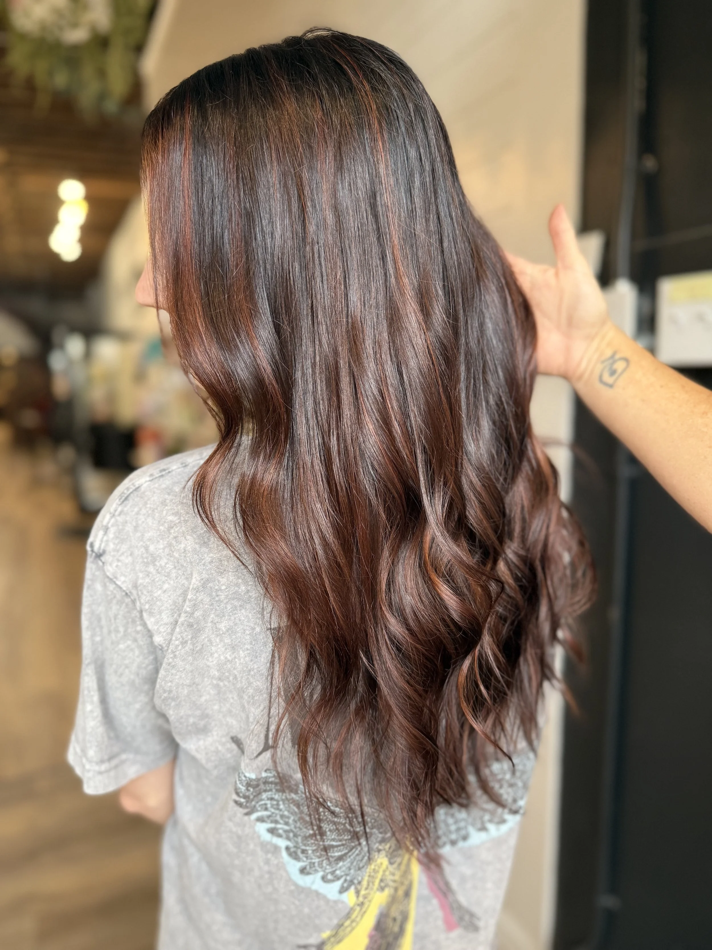 Woman with long, wavy, dark brown hair with reddish highlights, seen from the back, in a salon setting.