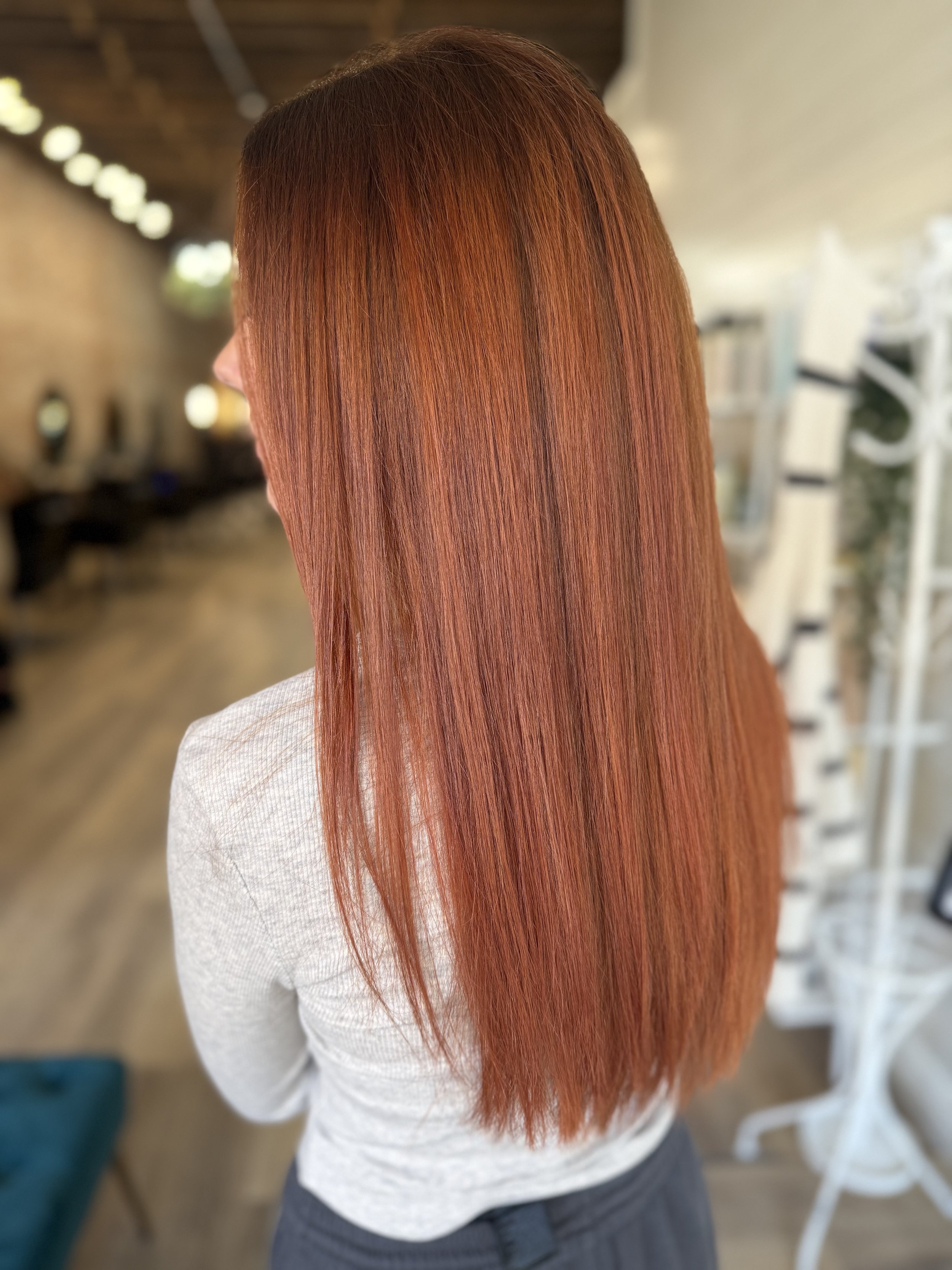 Close-up of a woman with long, straight, red hair wearing a white sweater and dark pants, standing indoors in a salon or similar setting.