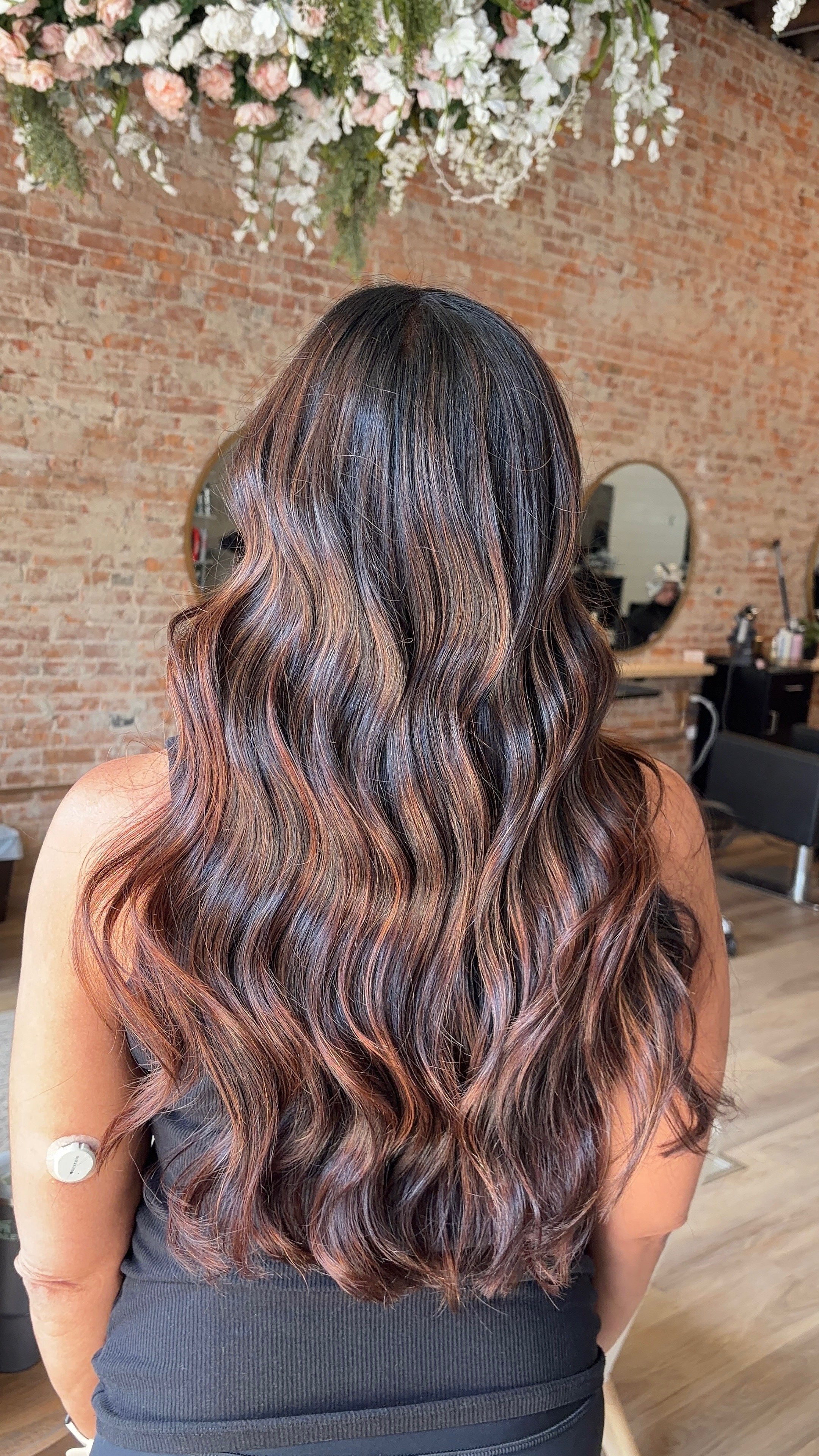 A woman with long, wavy hair showing a gradient of colors from dark brown at the roots to copper and rose gold highlights towards the ends, in a salon with a brick wall and floral decoration in the background.