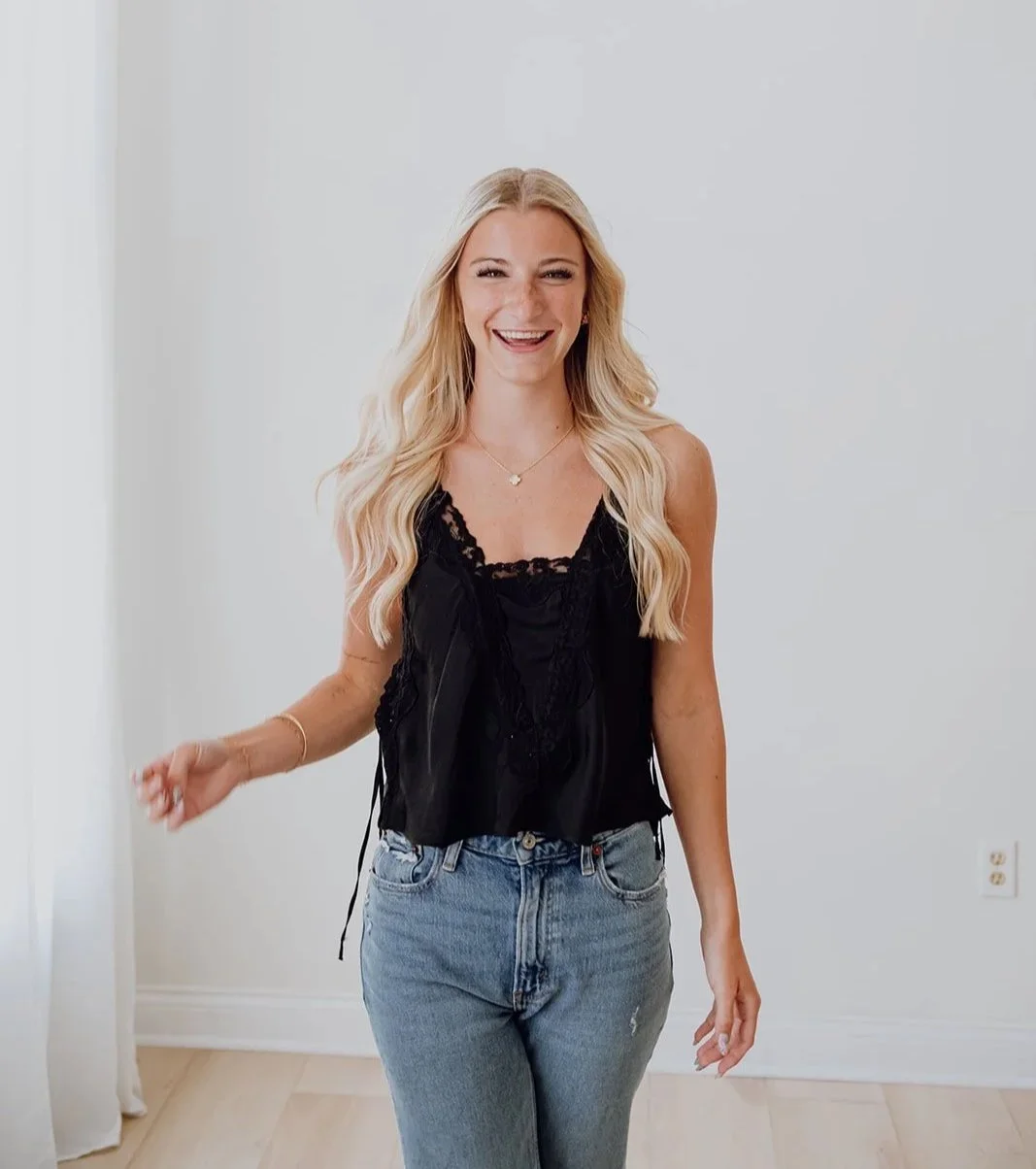 A smiling woman with long blonde hair, wearing a black sleeveless top with lace and faded jeans, standing in a bright, minimalist room.