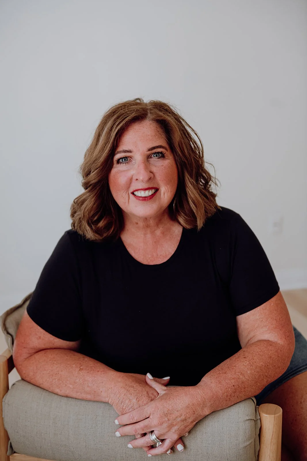 A woman with shoulder-length wavy brown hair, blue eyes, wearing a black t-shirt, smiling, seated on a beige chair with hands resting on her lap, in front of a plain white wall.