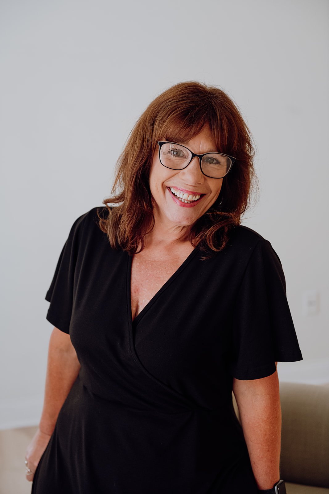 Smiling woman with brown hair, glasses, and a black dress, standing in a room with white walls.
