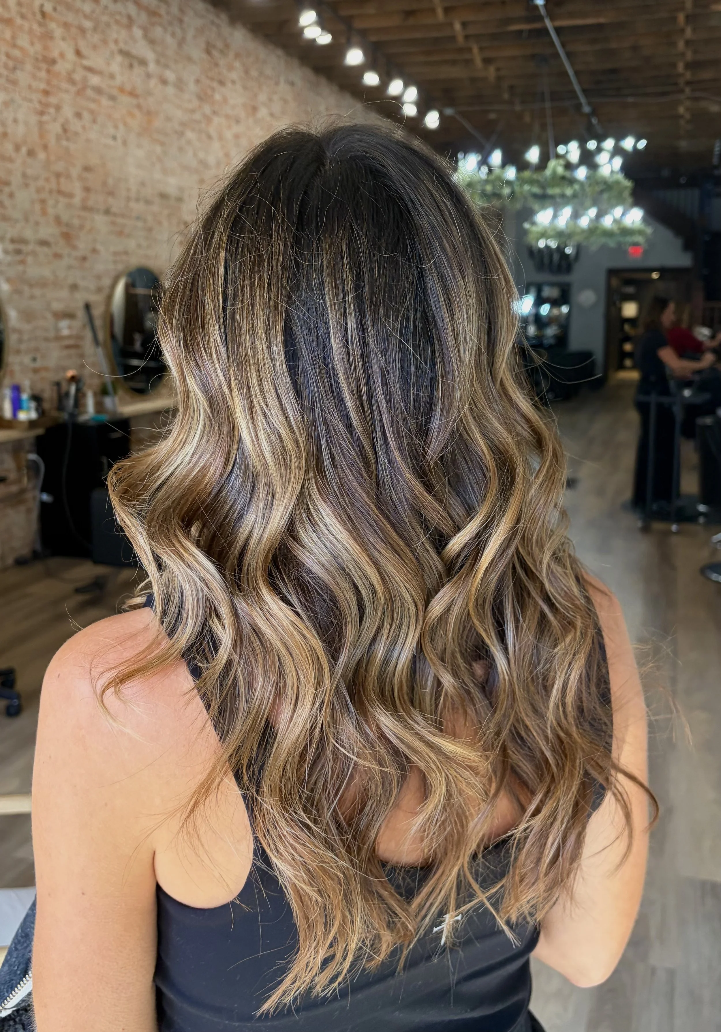 Woman with long, curled, ombre hair with darker roots transitioning to light brown and blonde tips, wearing a black sleeveless top in a salon.