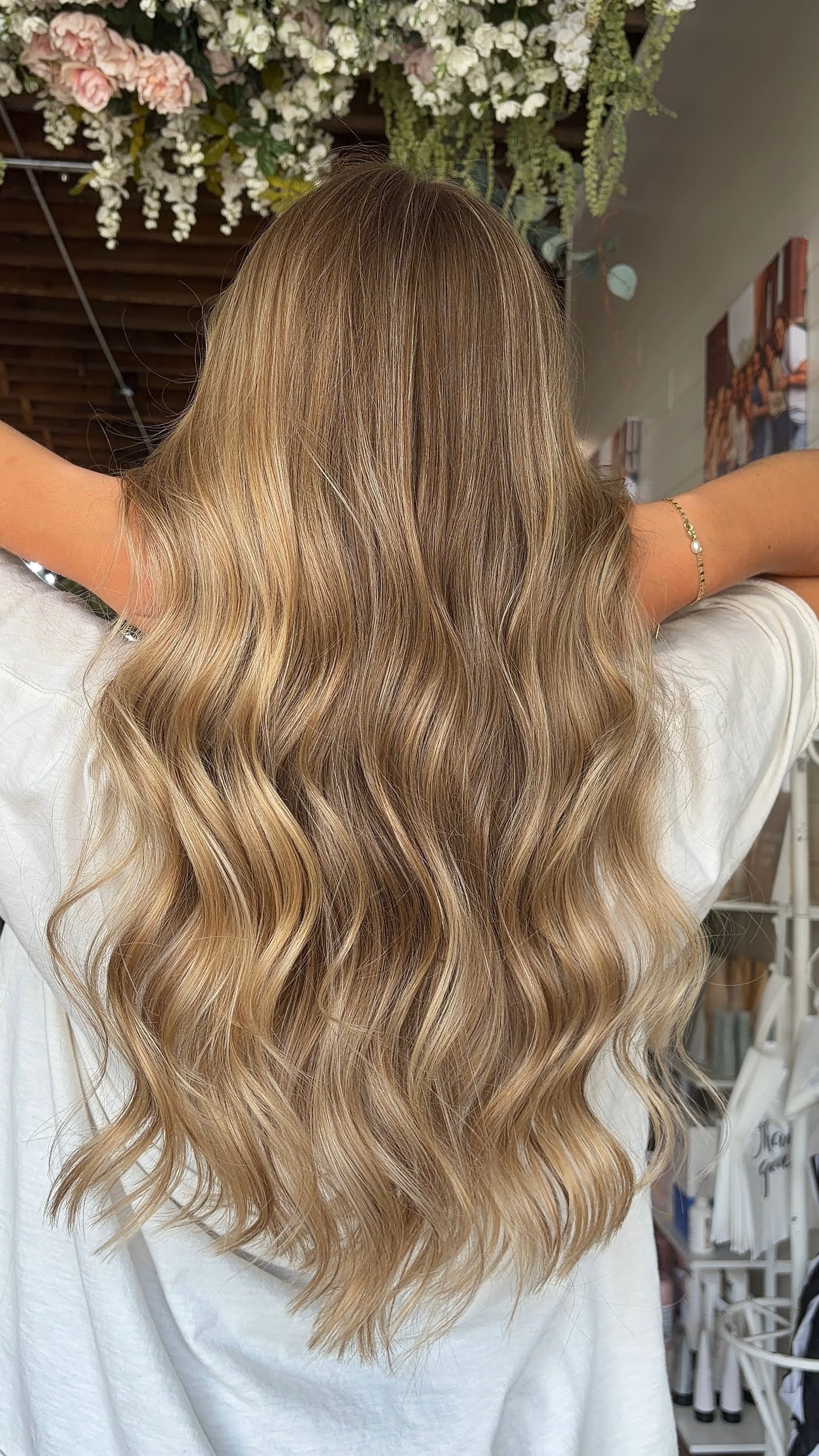 Person with long, wavy blonde hair standing with arms raised, back turned, in a salon or similar setting with floral decorations and salon equipment visible.