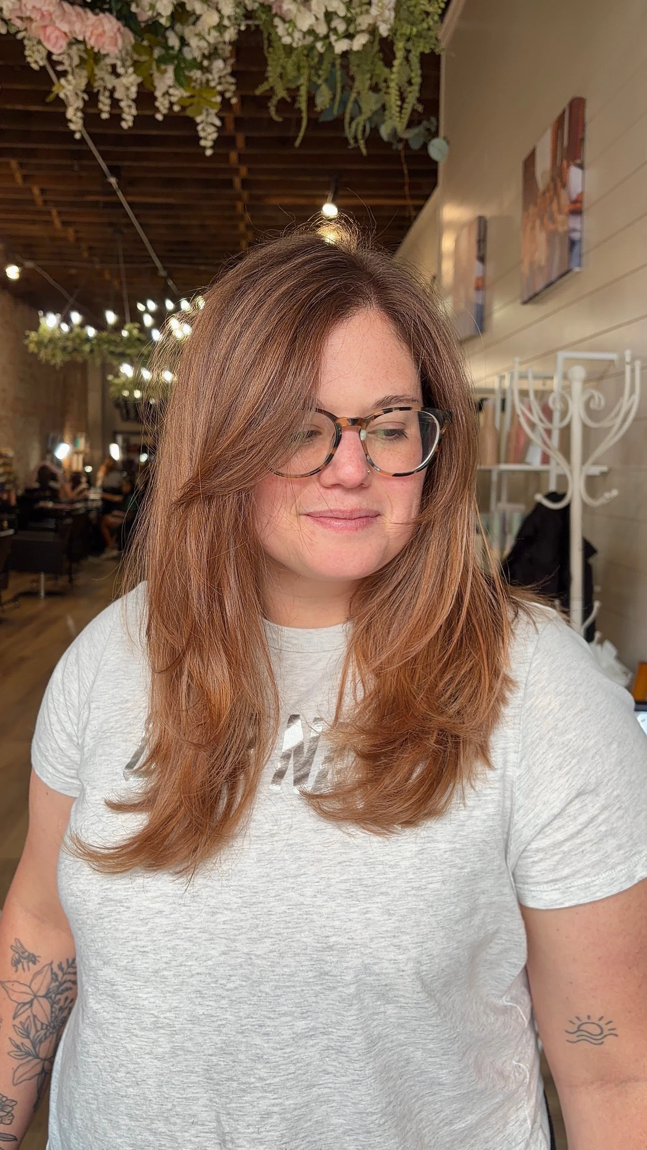 A woman with long red hair and glasses, wearing a light gray t-shirt, smiling slightly while looking down, in a cafe with wooden ceiling, hanging lights, and floral decorations.