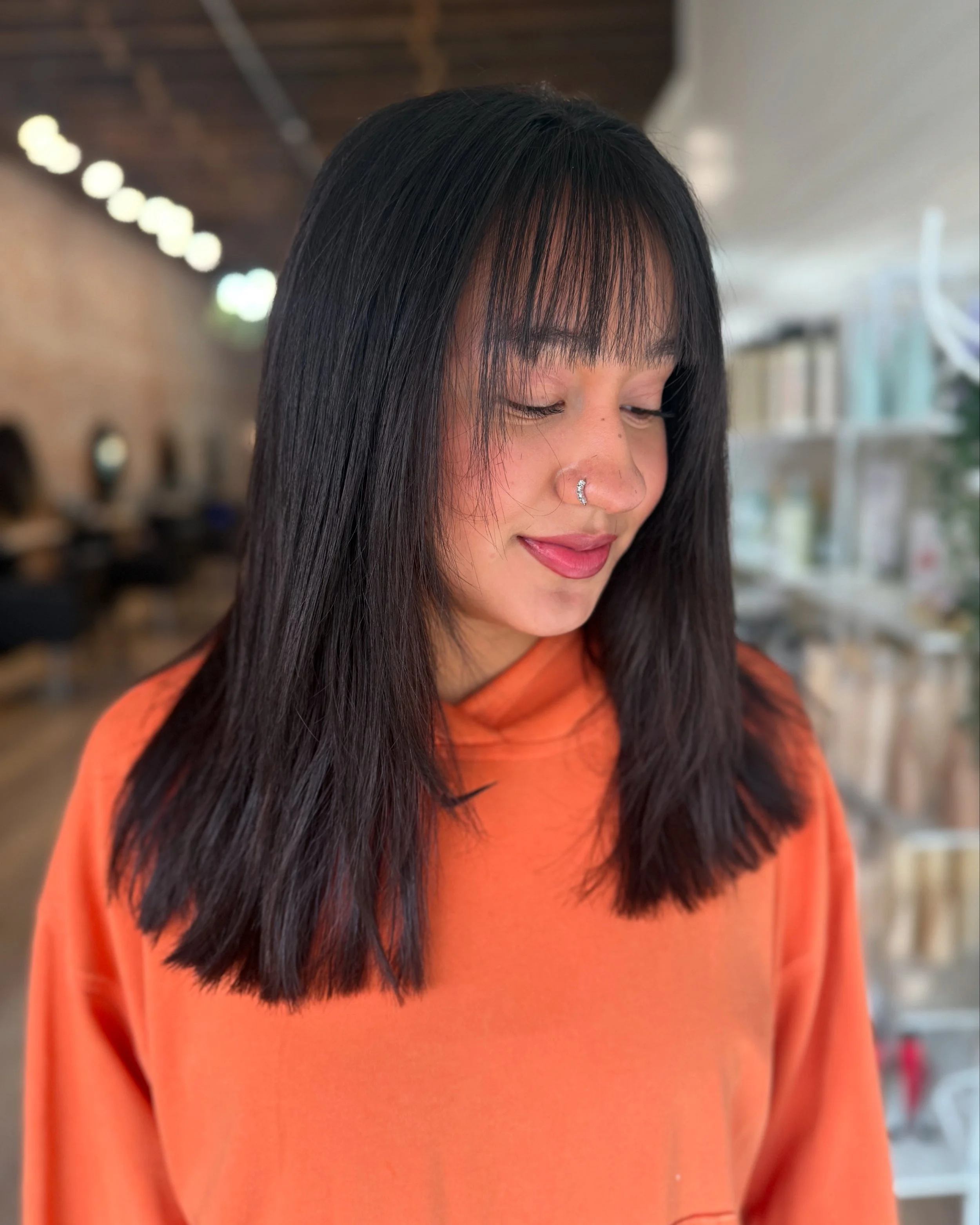 Close-up of a woman with straight black hair, wearing an orange top, inside a store or cafe with blurred background.