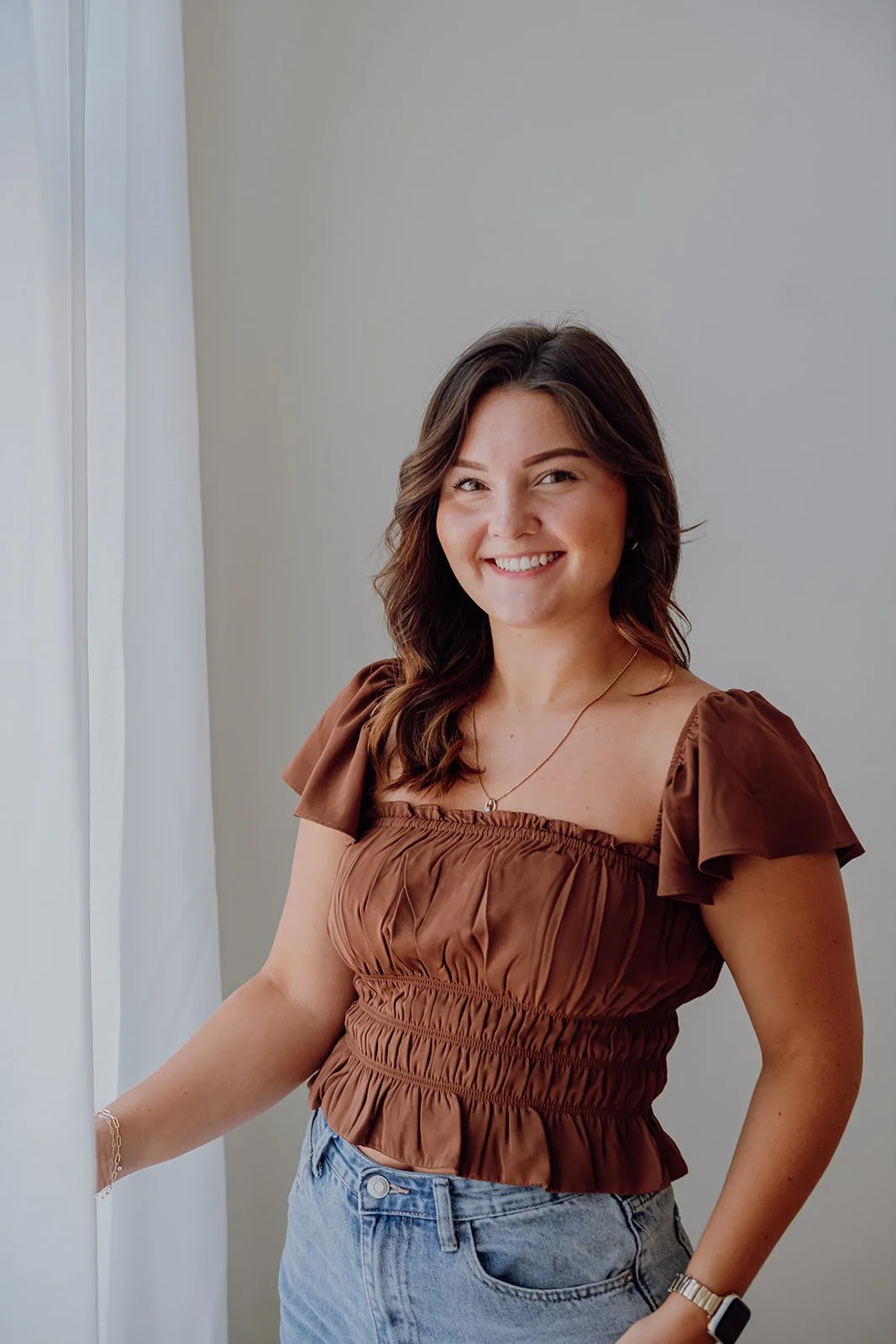 A young woman with shoulder-length brown hair smiling and standing by a white curtain, wearing a brown gathered top and light blue jeans.