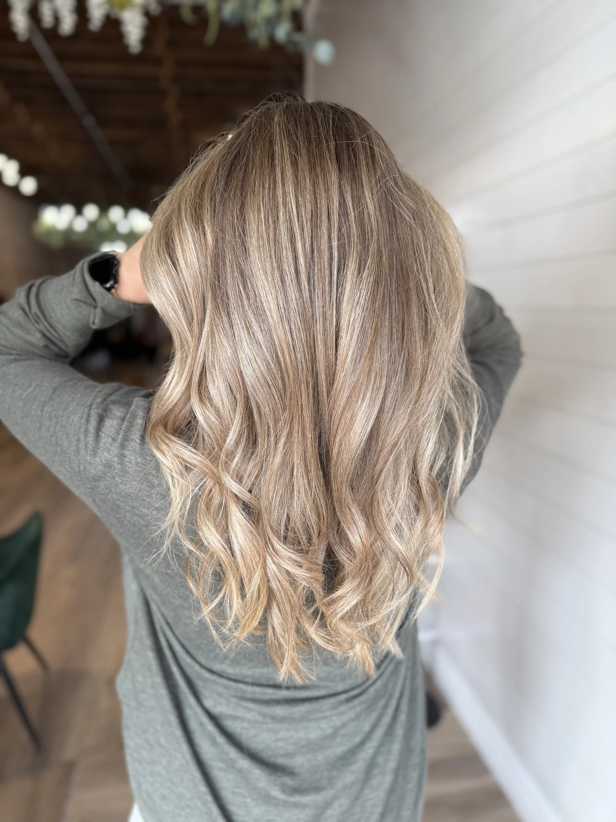 Top view of a woman's long, wavy blonde hair with subtle highlights, wearing a gray sweater, in a room with a white wall and wooden ceiling.
