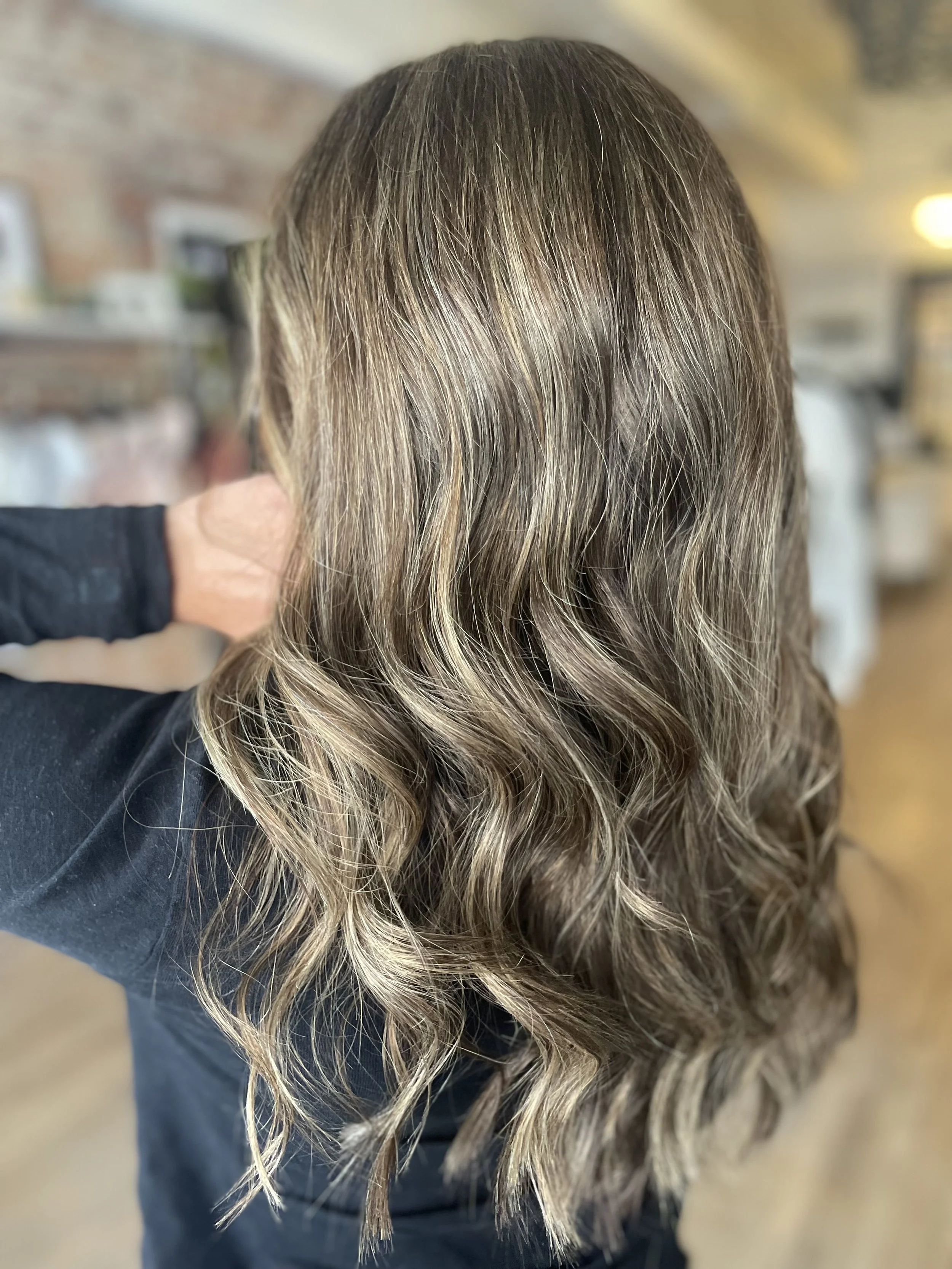 Close-up of a person's brown hair with blonde highlights, styled with loose curls, in an indoor setting.