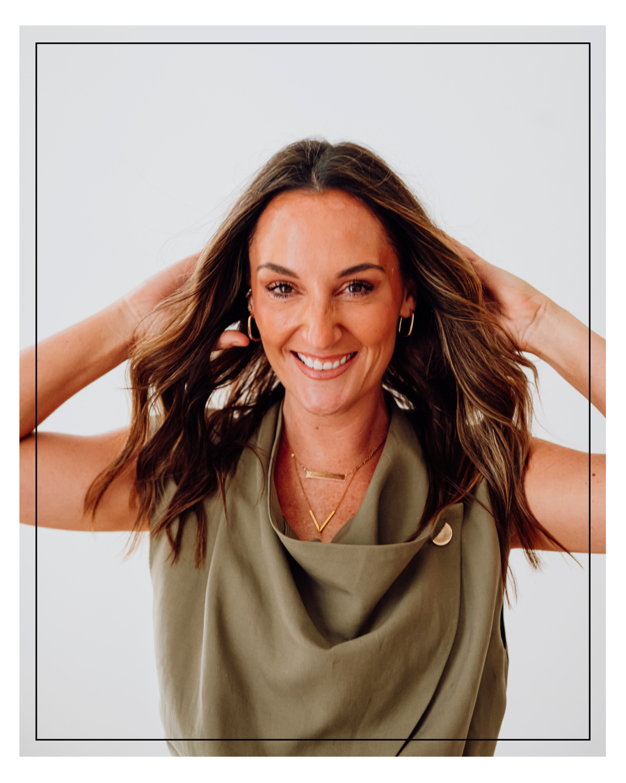 Smiling woman with wavy brown hair wearing gold hoop earrings and layered necklaces, posing with hands behind her head against a white background.