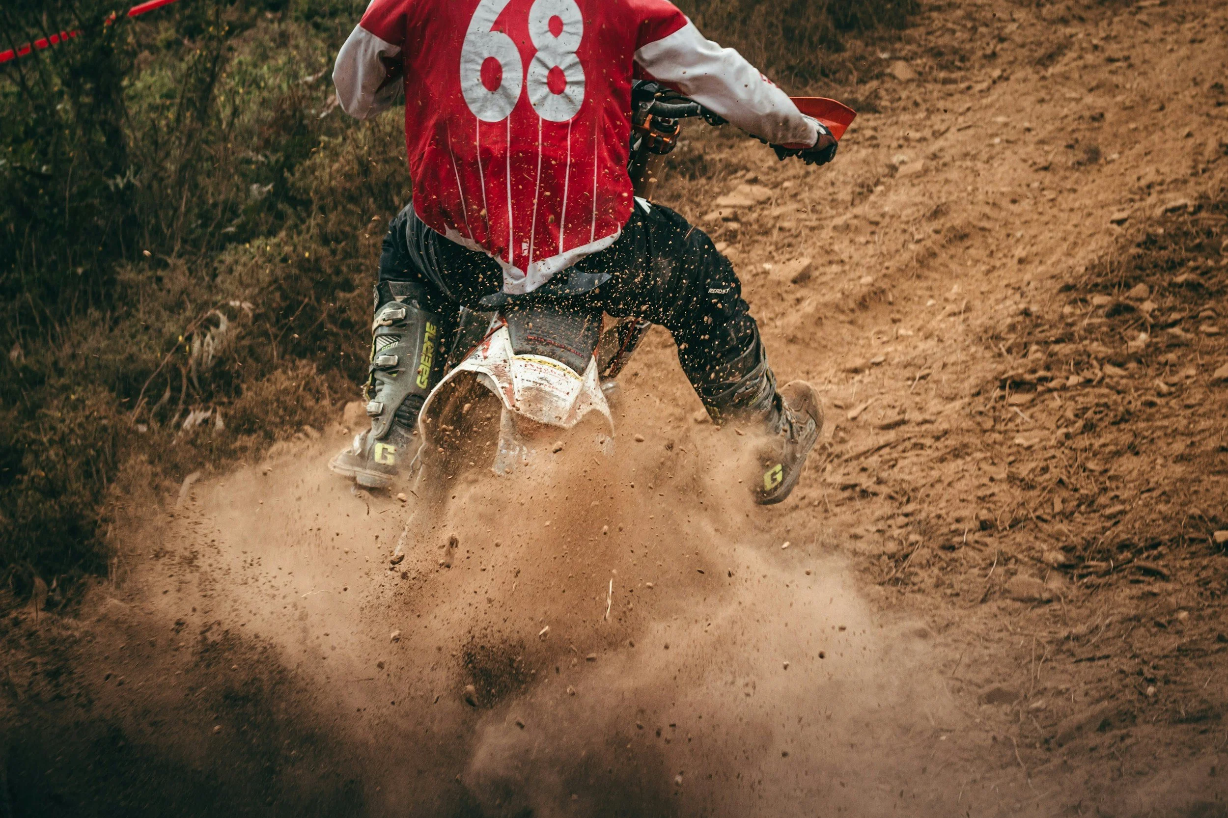 A person riding a dirt bike on a dirt trail, wearing black protective gear and a red and white jersey with large white numbers.