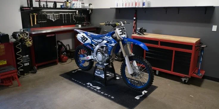 Blue and white motocross dirt bike with number 38 on the front, standing on a black mat in a garage or workshop.