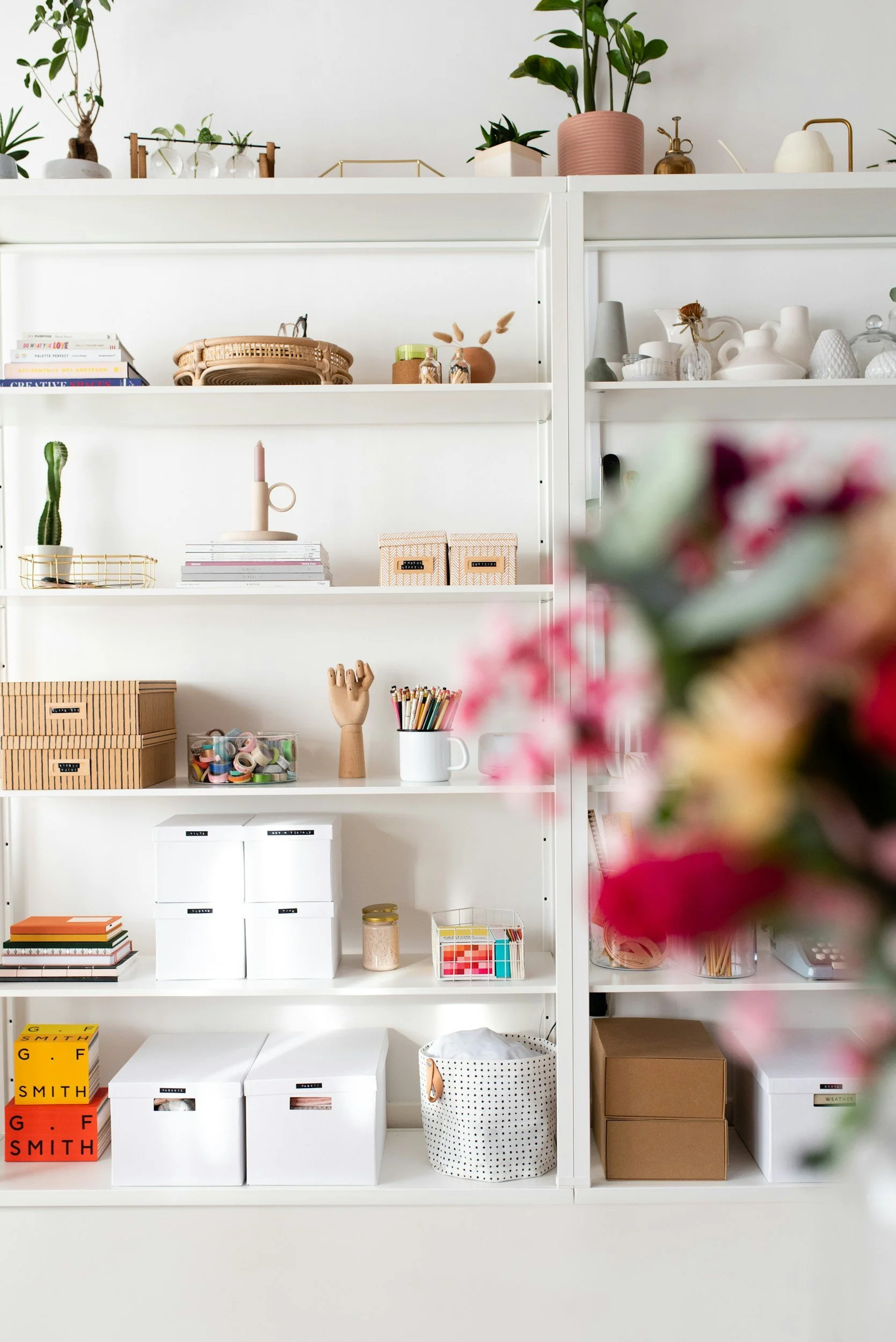 A white shelving unit filled with decorative and functional items such as books, storage boxes, plants, vases, candles, and organizing containers, with a blurred pink flower arrangement in the foreground.