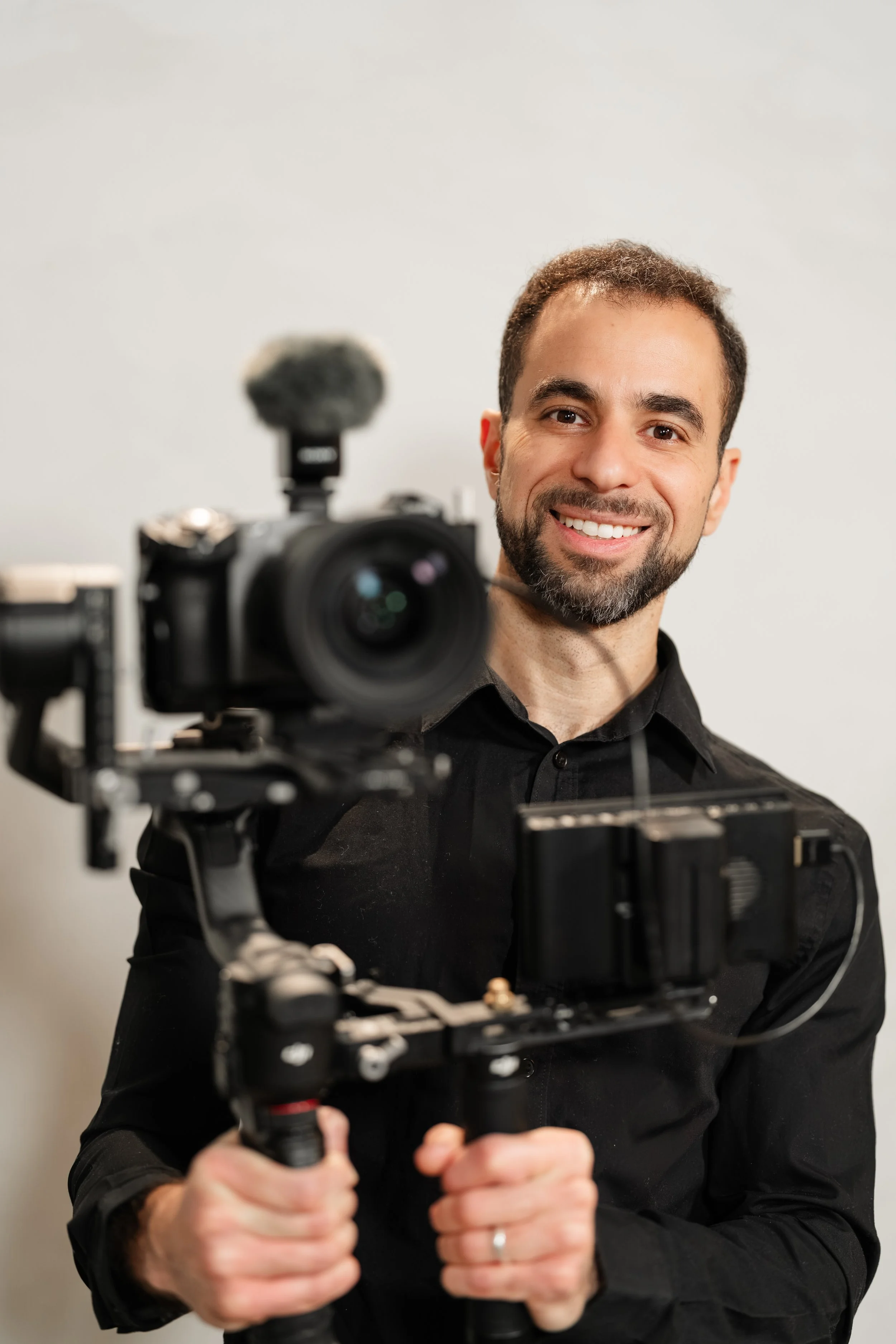 A smiling man with short brown hair and a beard is holding a professional camera mounted on a stabilizer. He is wearing a black shirt and standing in front of a plain light-colored background.