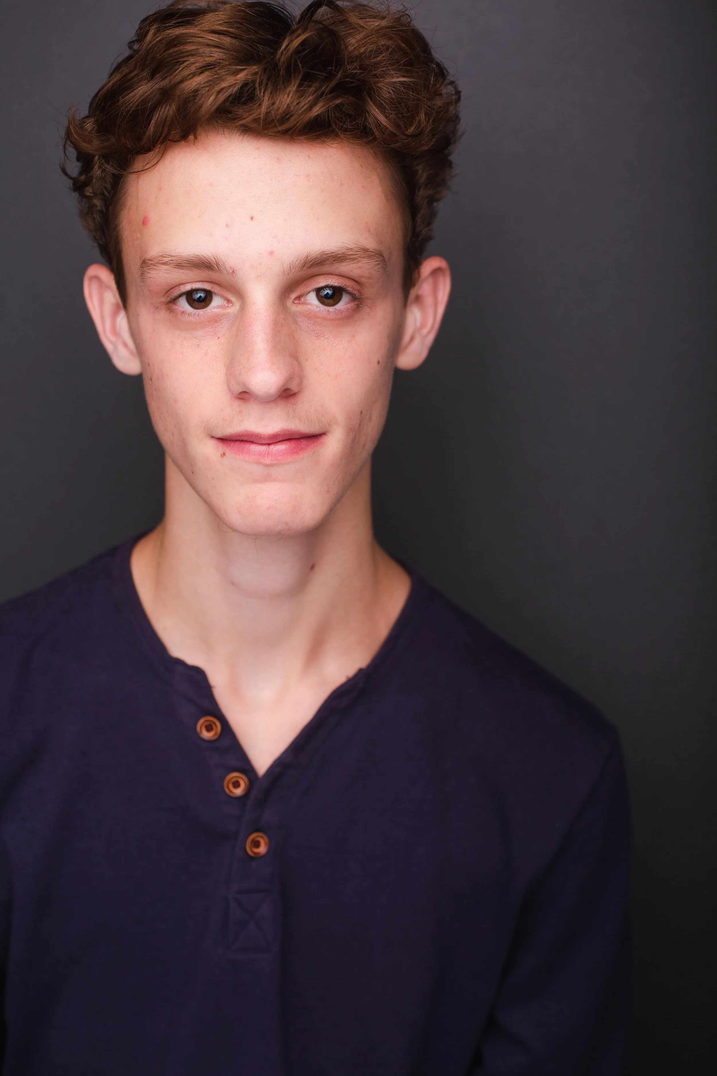 A young man with curly brown hair and fair skin posing against a dark background, wearing a navy blue shirt with brown buttons.