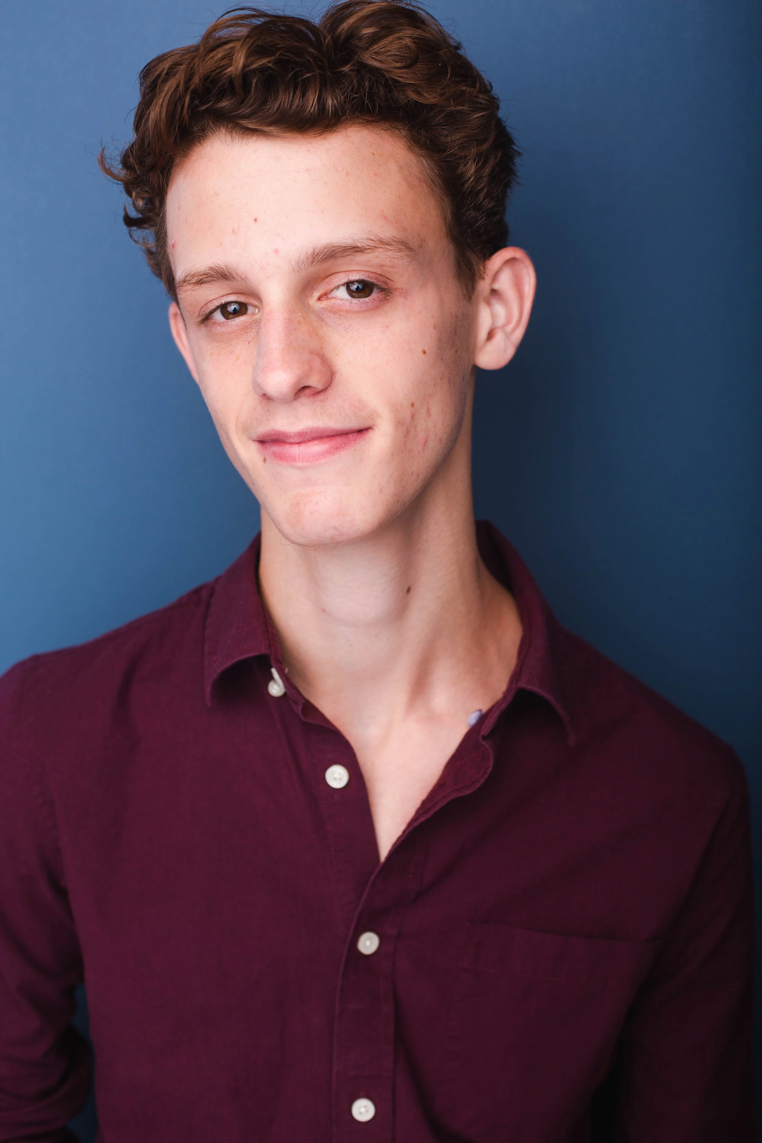 A young man with light skin, wavy brown hair, and a slight smile, wearing a maroon button-up shirt, standing against a blue background.