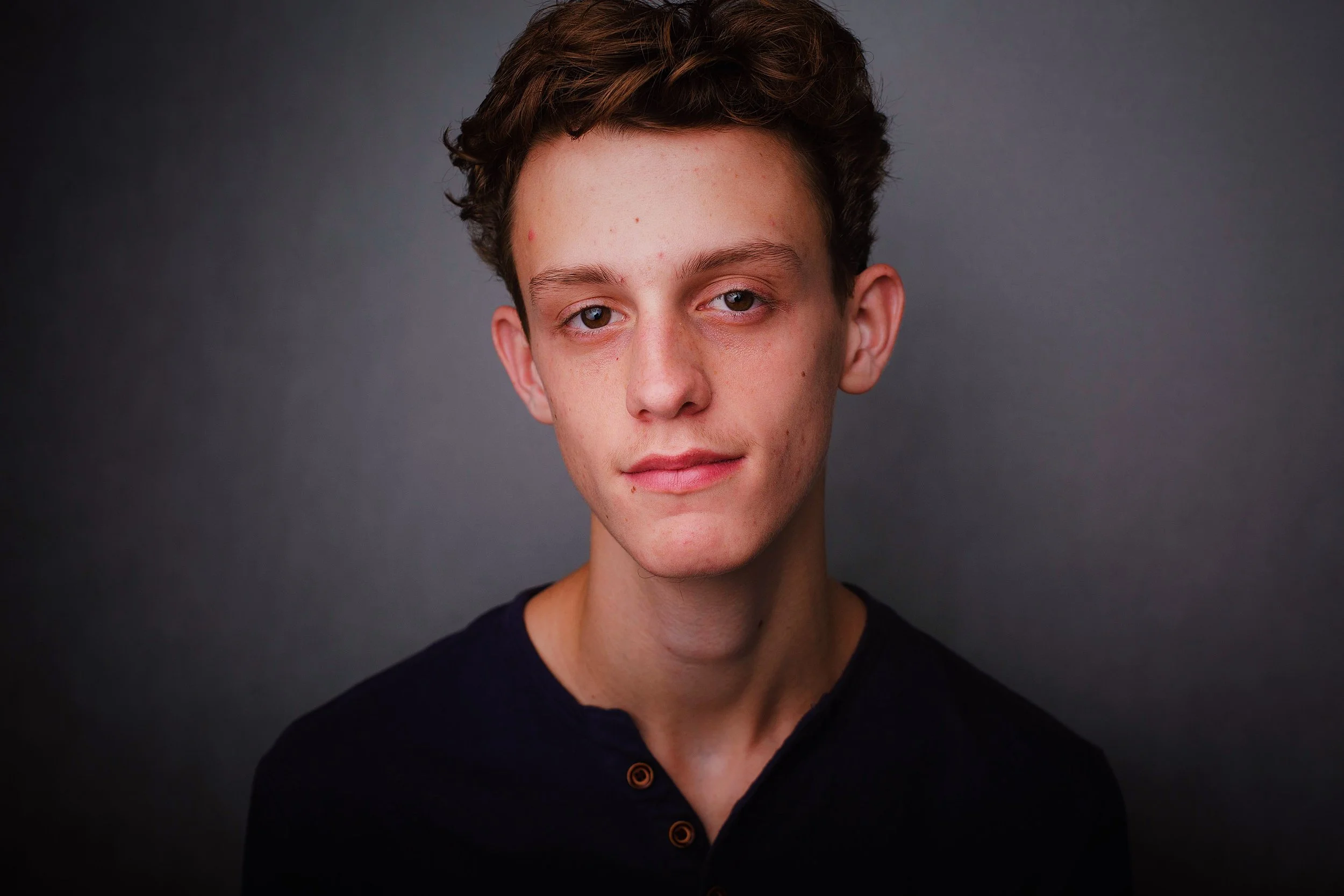 Portrait of a young man with short, curly brown hair, wearing a black shirt, against a dark background.