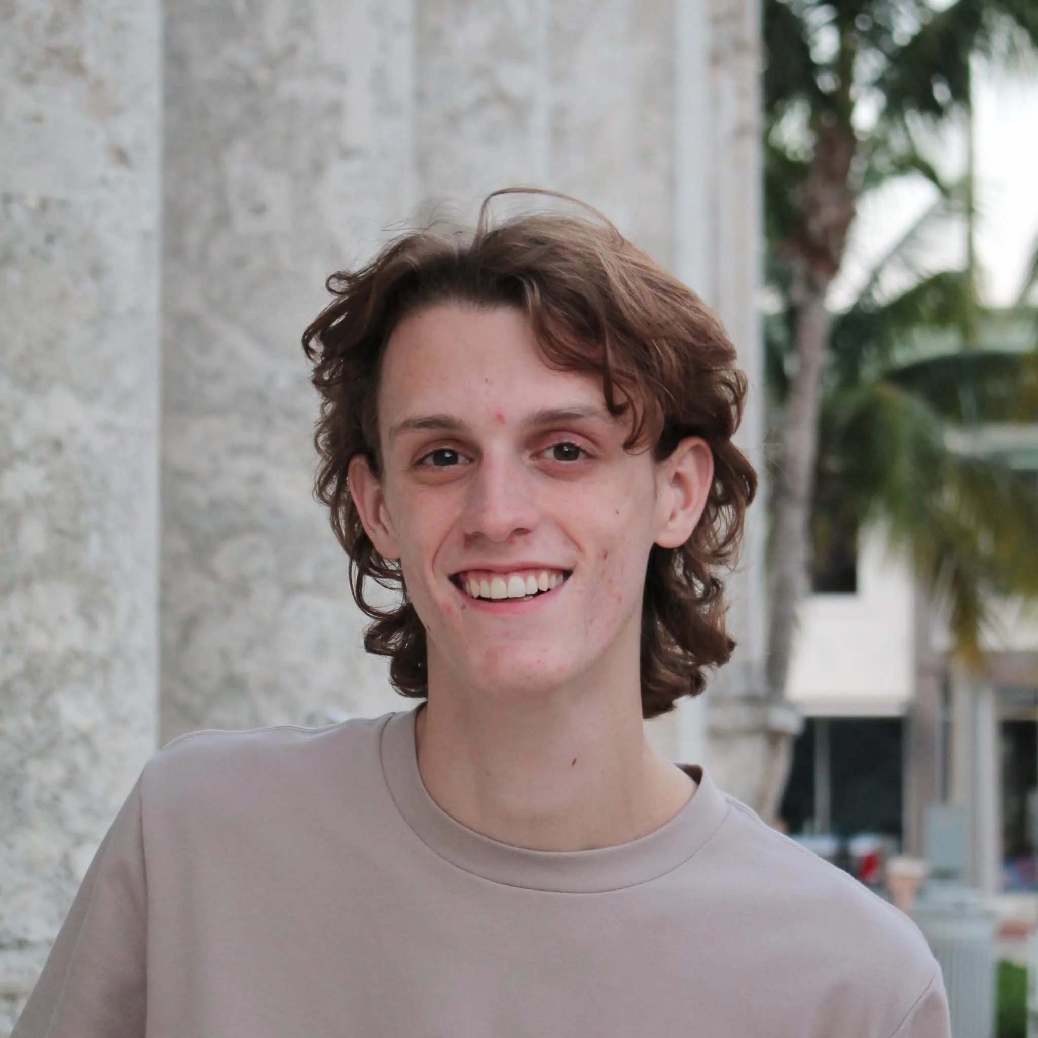A young man with shoulder-length wavy brown hair smiling outdoors in front of a stone building and palm trees.