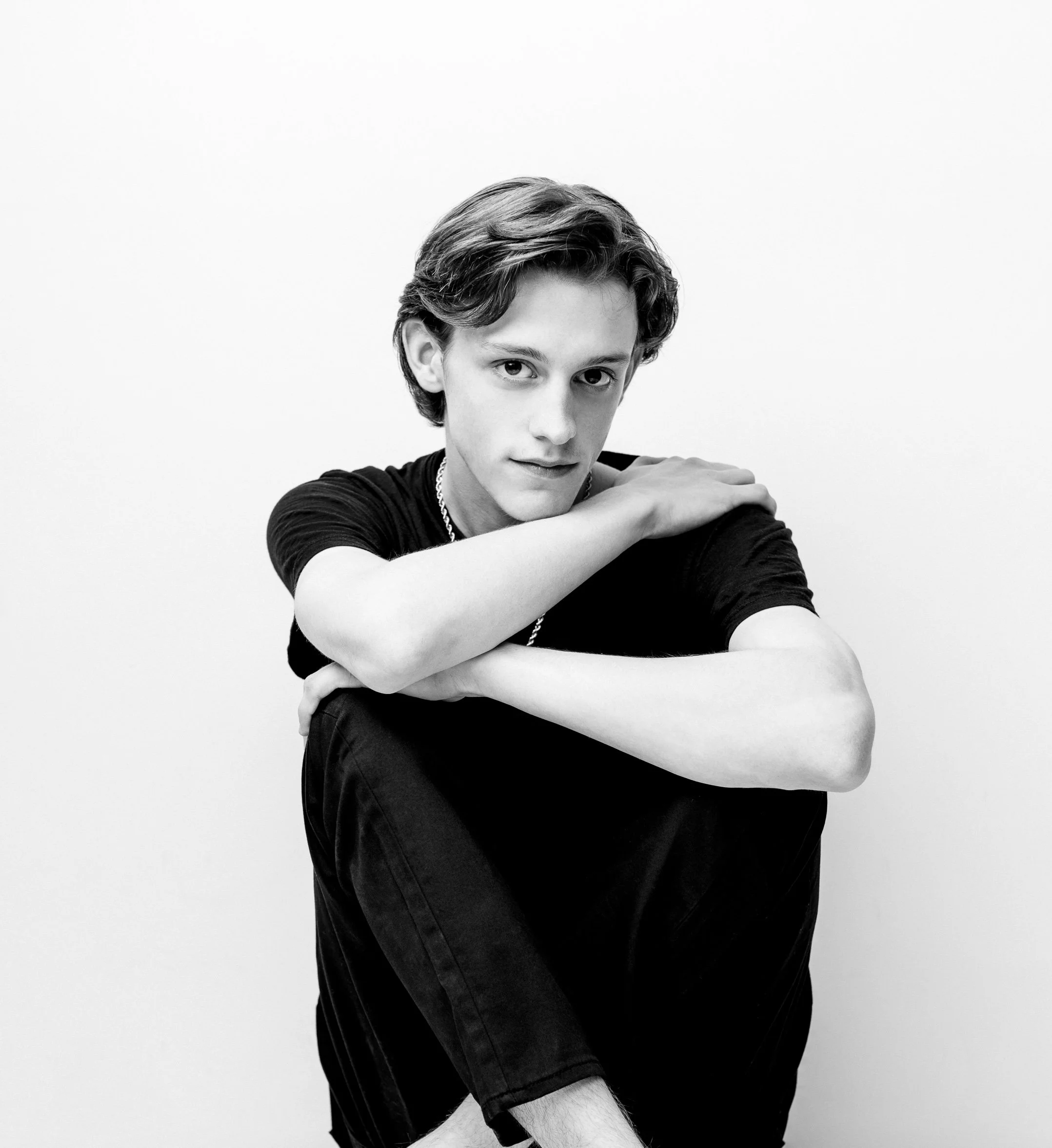 A black and white portrait of a young man with wavy hair, sitting with his arms crossed over his knees, wearing a black T-shirt and a chain necklace, against a plain white background.
