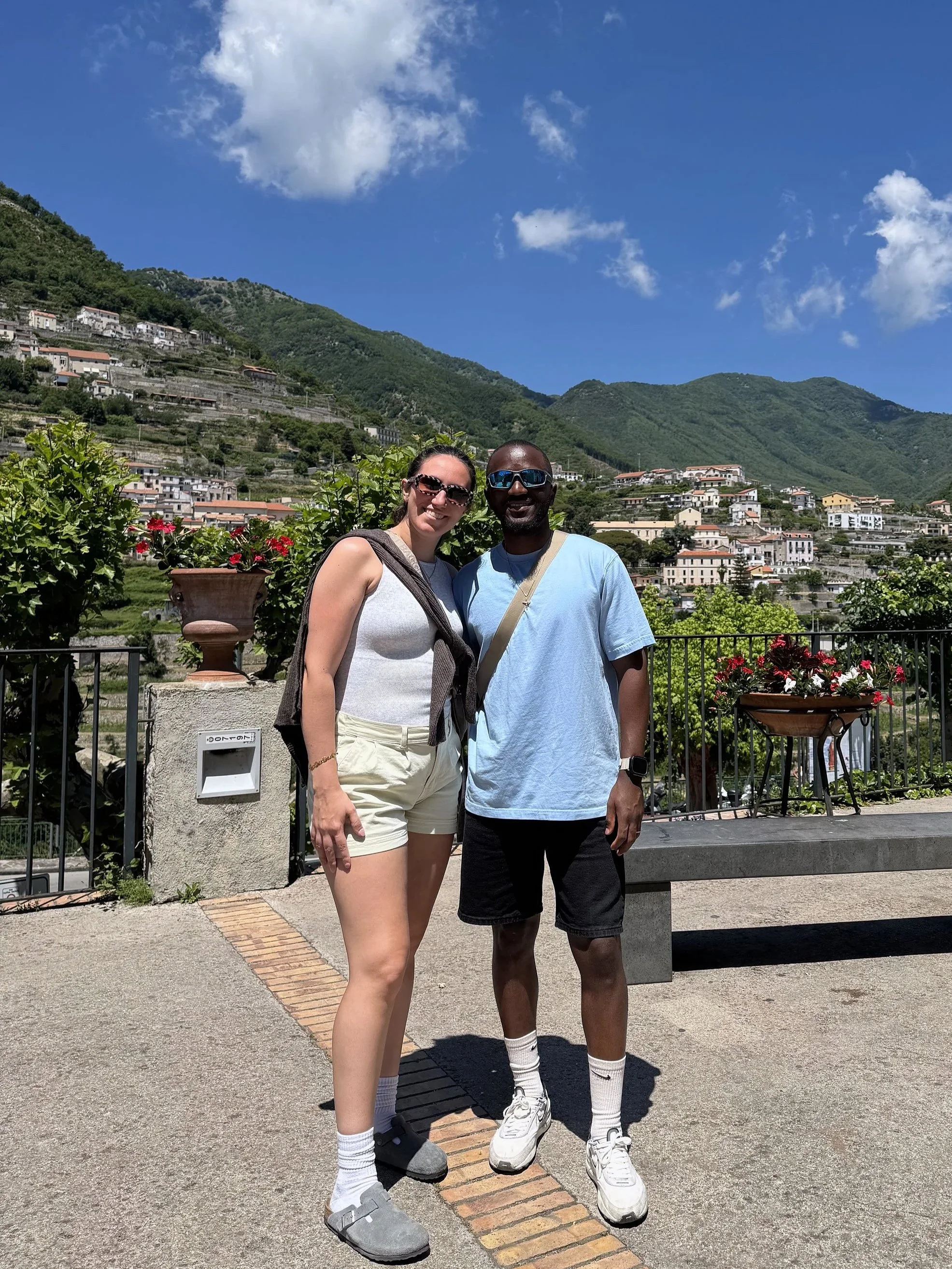Two people smiling and posing for a photo outdoors with a mountainous landscape in the background. One woman, wearing a sleeveless top, shorts, gray Crocs, and sunglasses, is standing next to a man in a light blue t-shirt, black shorts, white sneakers, and sunglasses. There are potted plants with red and white flowers and a fence behind them.