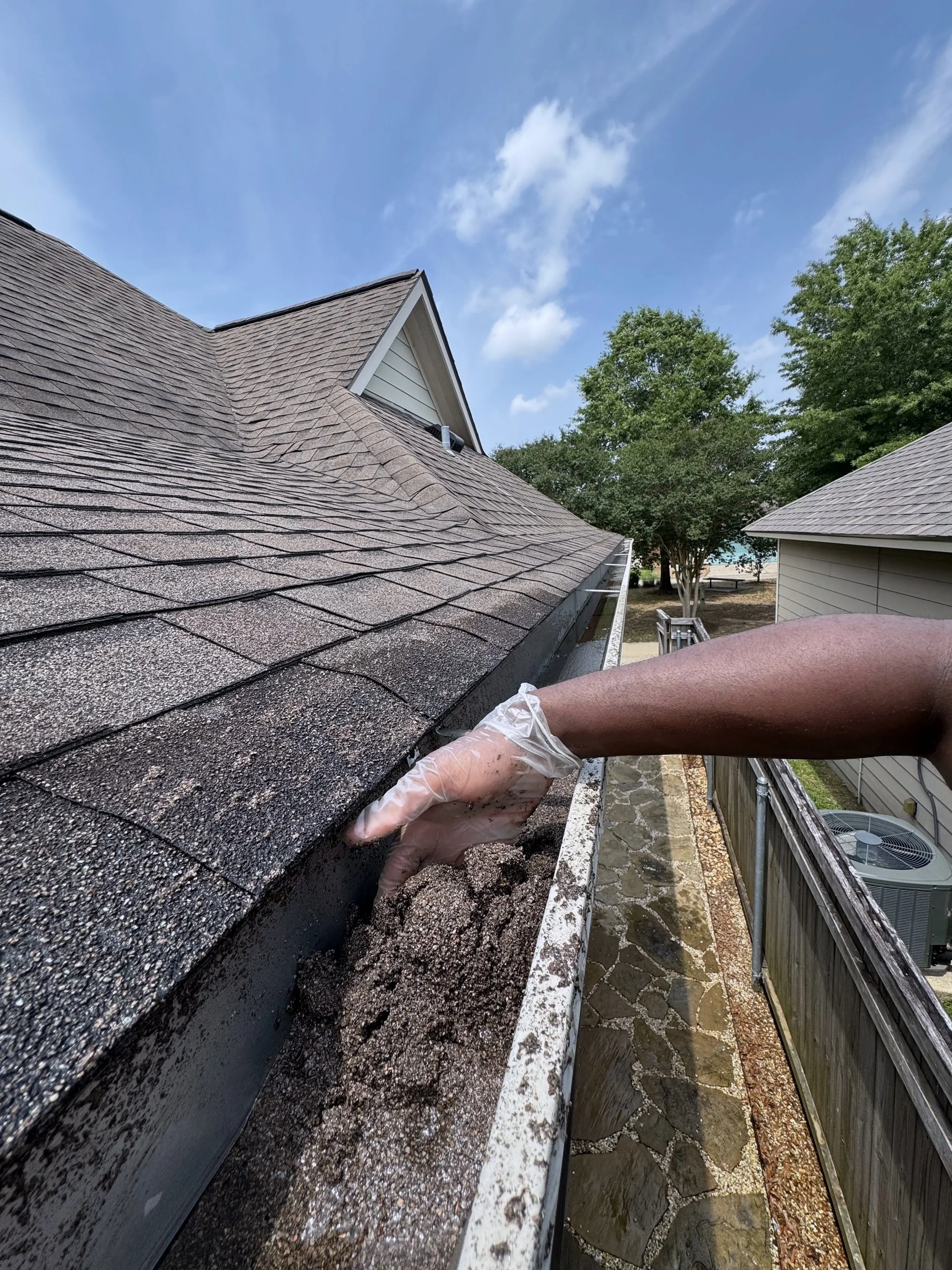 Worker cleaning or repairing a roof with shingles, wearing gloves and a plastic covering on the arm, with trees, sky, and other buildings in the background.