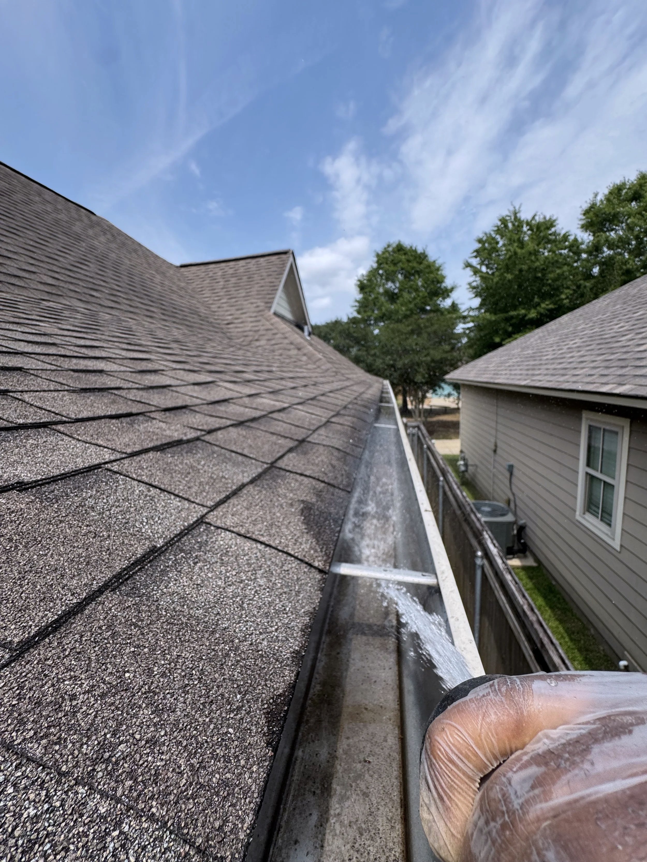 Close-up view of a person's knee and foot in protective plastic, cleaning or working on a roof with a hose, shingles, gutters, and neighboring houses in the background under a blue sky with clouds.