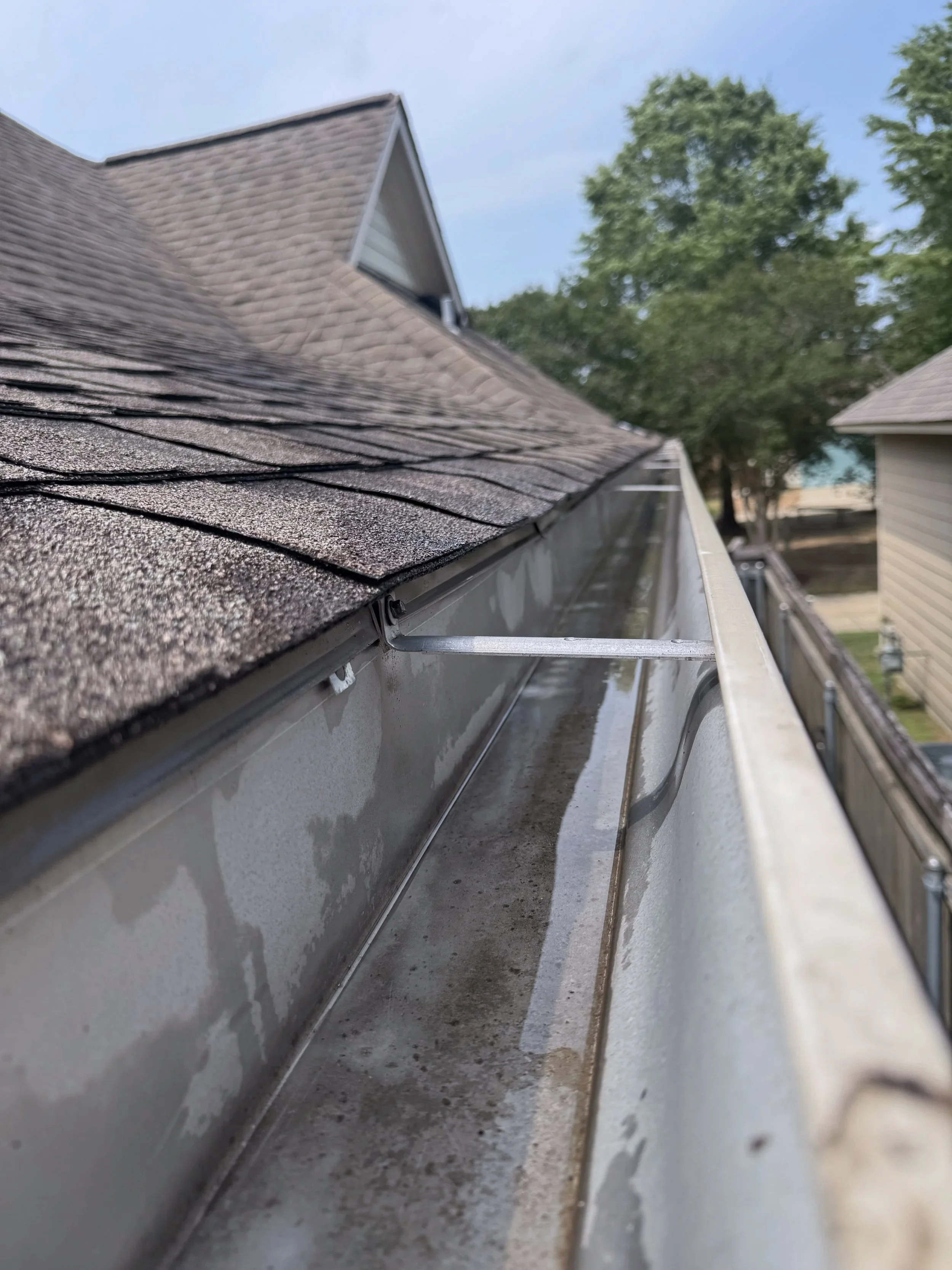Close-up of a house's roof with shingle roofing, gutter system, and a clear blue sky with trees in the background.