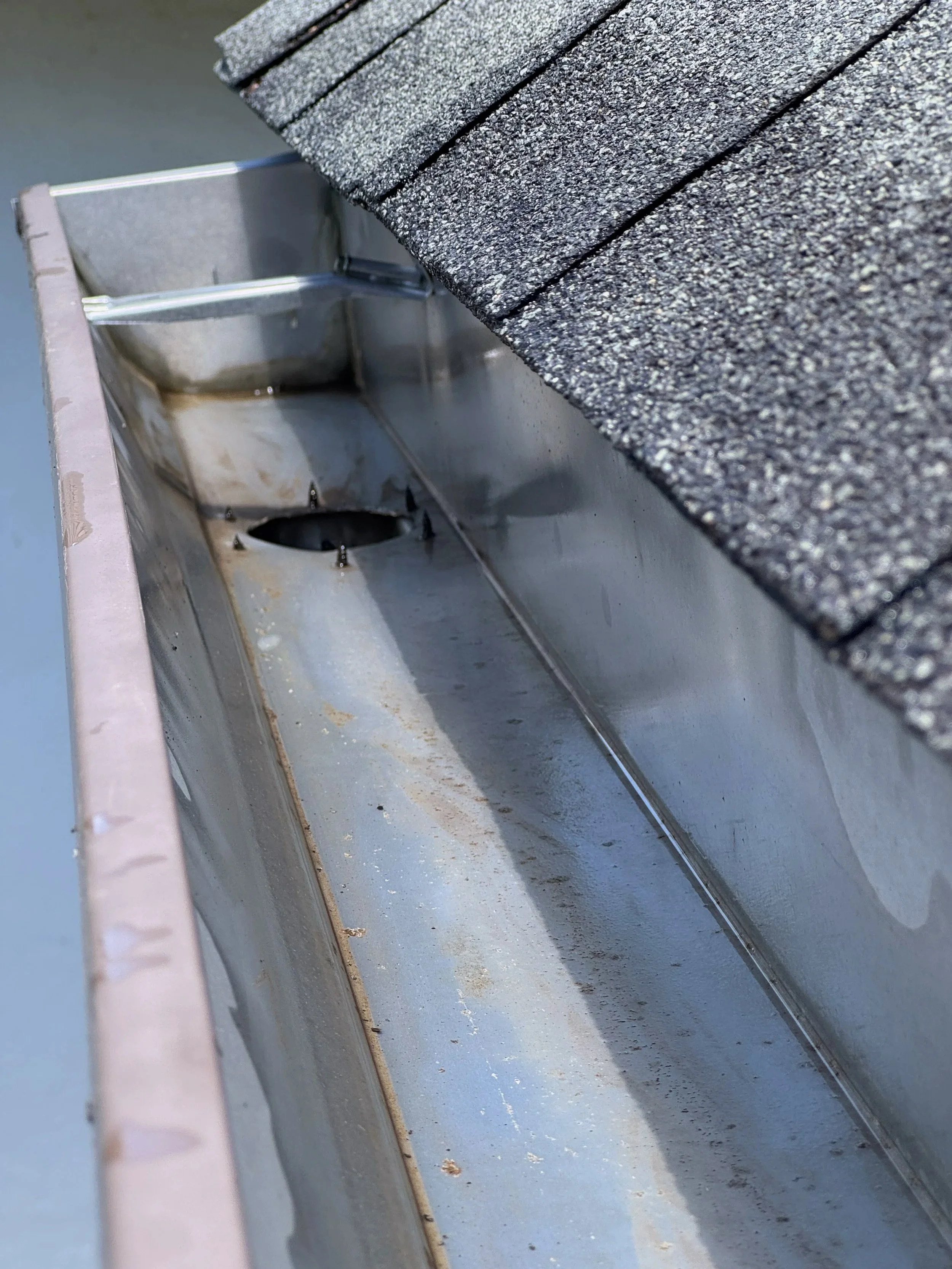 Close-up image of a metal gutter with a textured asphalt shingle roof above it. The gutter has some dirt and stains inside.