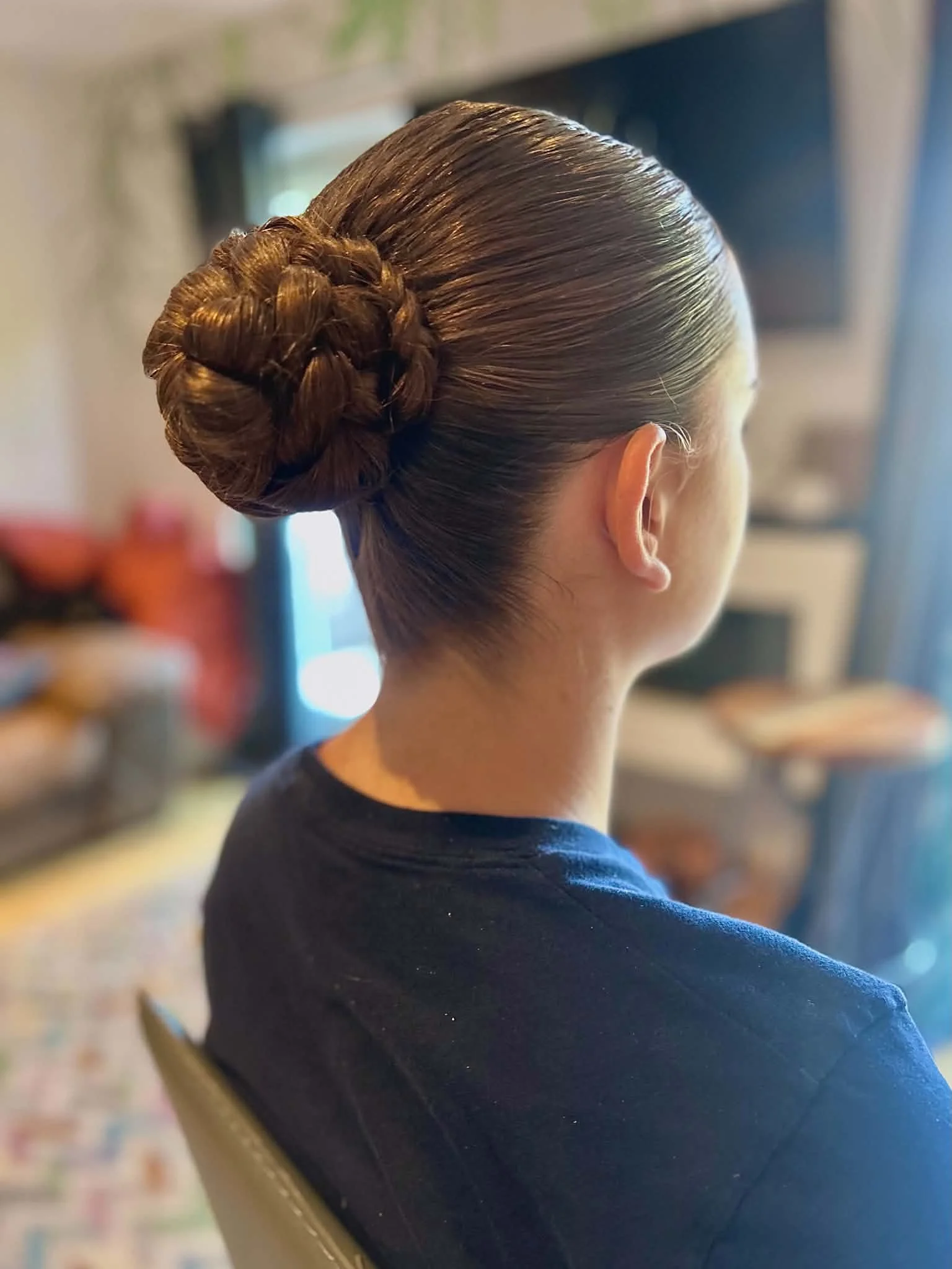 Close-up of a woman with neatly styled brown hair in a bun, wearing a dark blue shirt. The background appears to be a room with blurred furniture and decor.