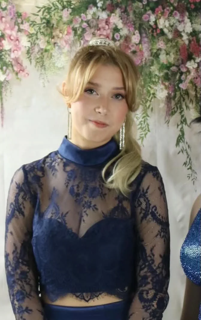A young woman with blonde hair styled in soft waves, wearing a delicate tiara and long earrings. She is dressed in a sheer navy blue lace top with floral patterns over a matching navy blue undergarment, standing in front of a floral backdrop with pin