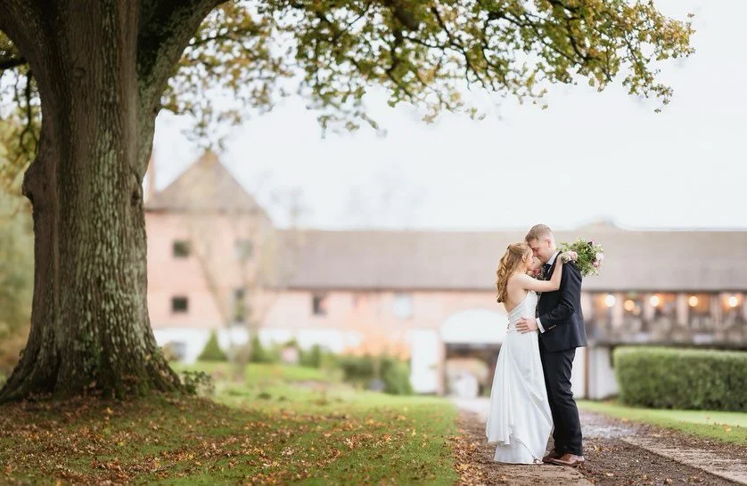 A bride and groom embrace in front of a large tree on their wedding day, with a rustic barn in the background.