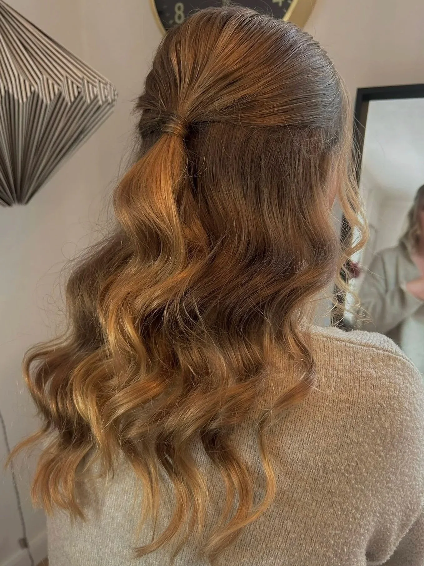 Woman with long, wavy, auburn hair styled in loose curls, wearing a beige top, standing in front of a mirror in a room with a clock on the wall.