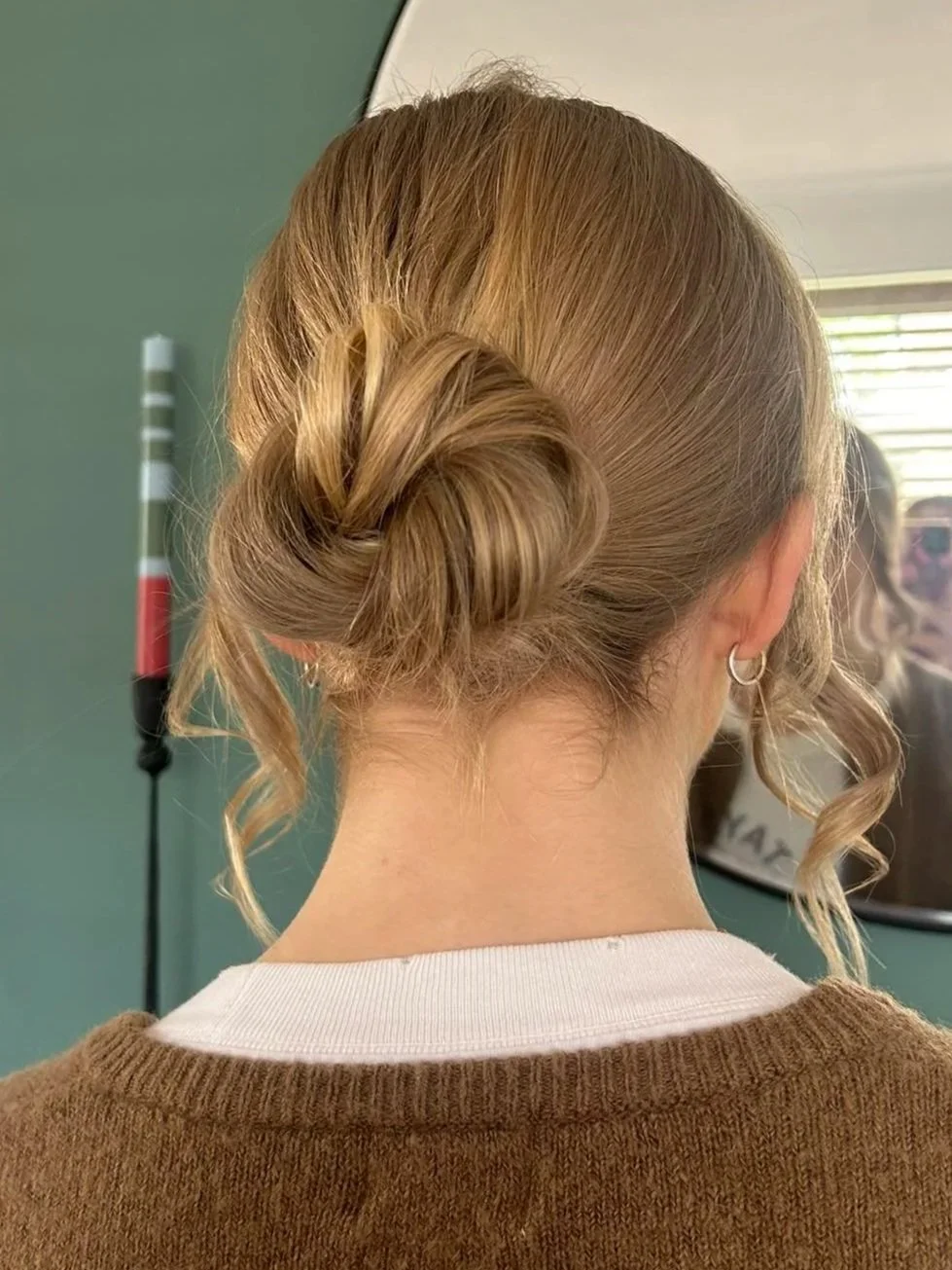 Back of a woman's head with blonde hair styled in a twisted bun, with some loose curls. She wears a white shirt under a brown sweater and a small hoop earring. In the background, a mirror shows her reflection and a green wall.