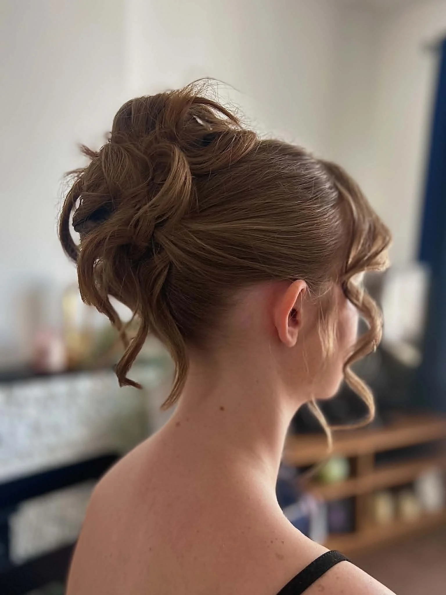 A woman with red hair styled in an elegant updo with curls, view from the side, wearing a black strap on her shoulder, in an indoor setting.