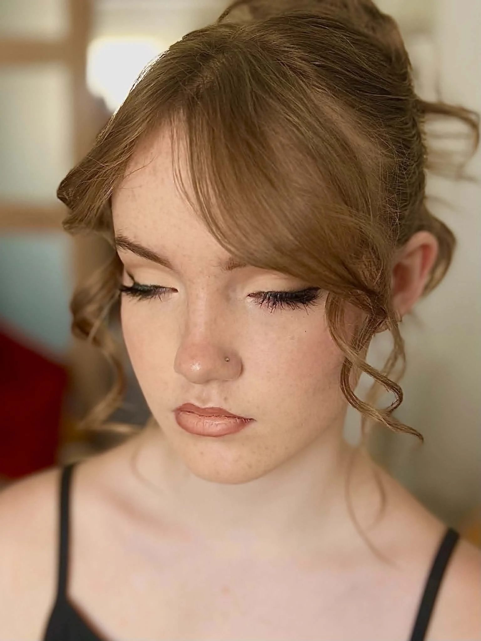 Close-up of a woman with auburn hair styled in loose curls and a side-swept bang, with eyes closed, with softy glam makeup with soft cat eye eyeliner.